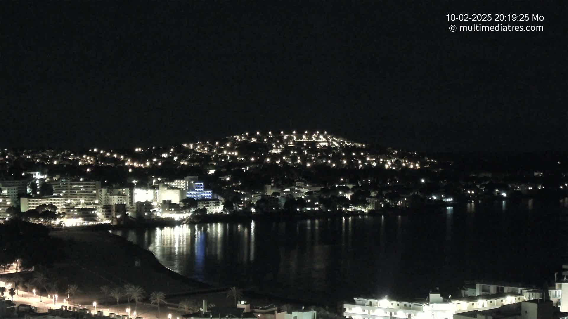 A nighttime view of a coastal town illuminated by lights, reflecting on a calm body of water.
