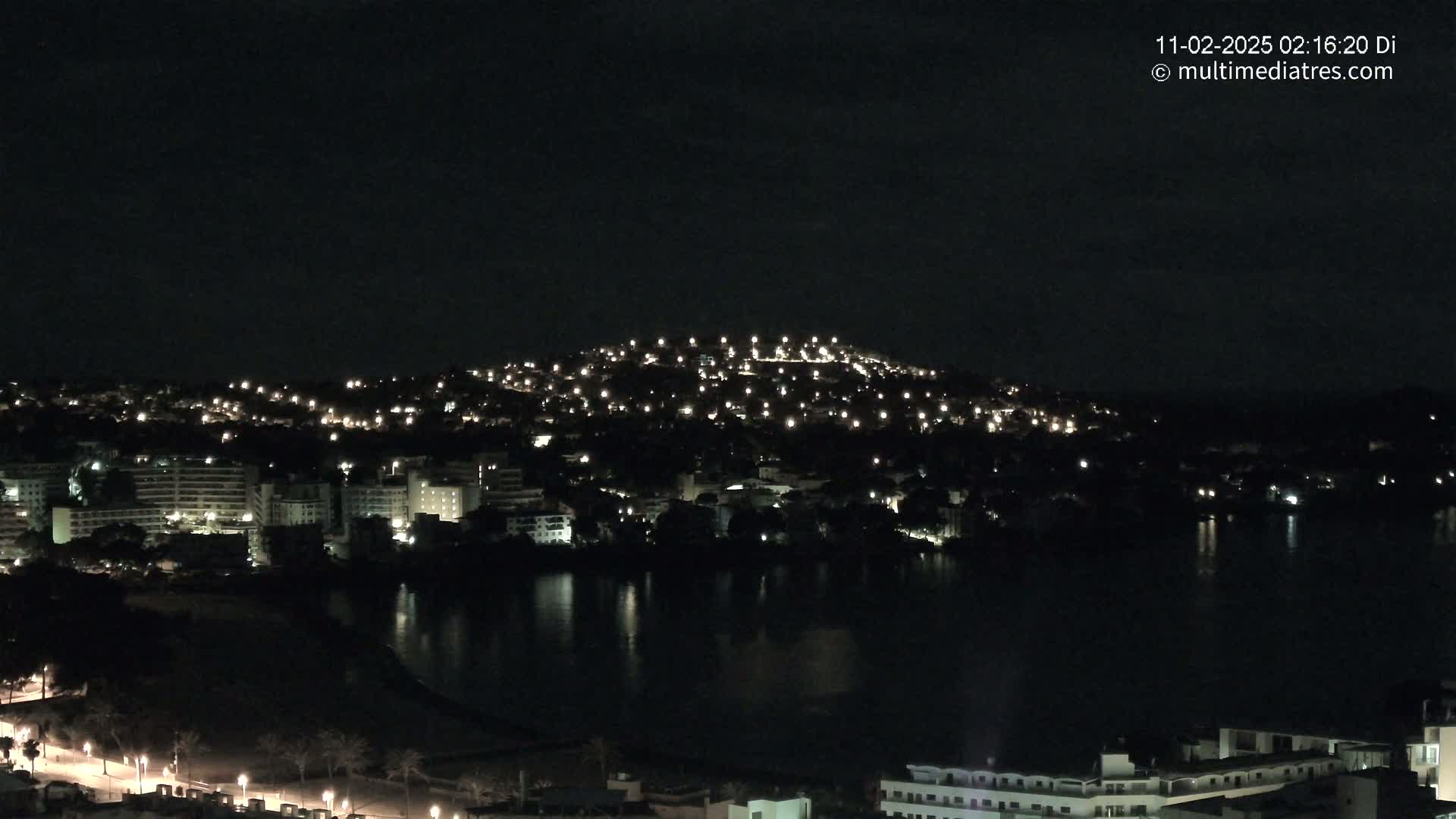 A nighttime view of a city and coastline illuminated by numerous lights, reflecting on a dark body of water under a clear, dark sky.