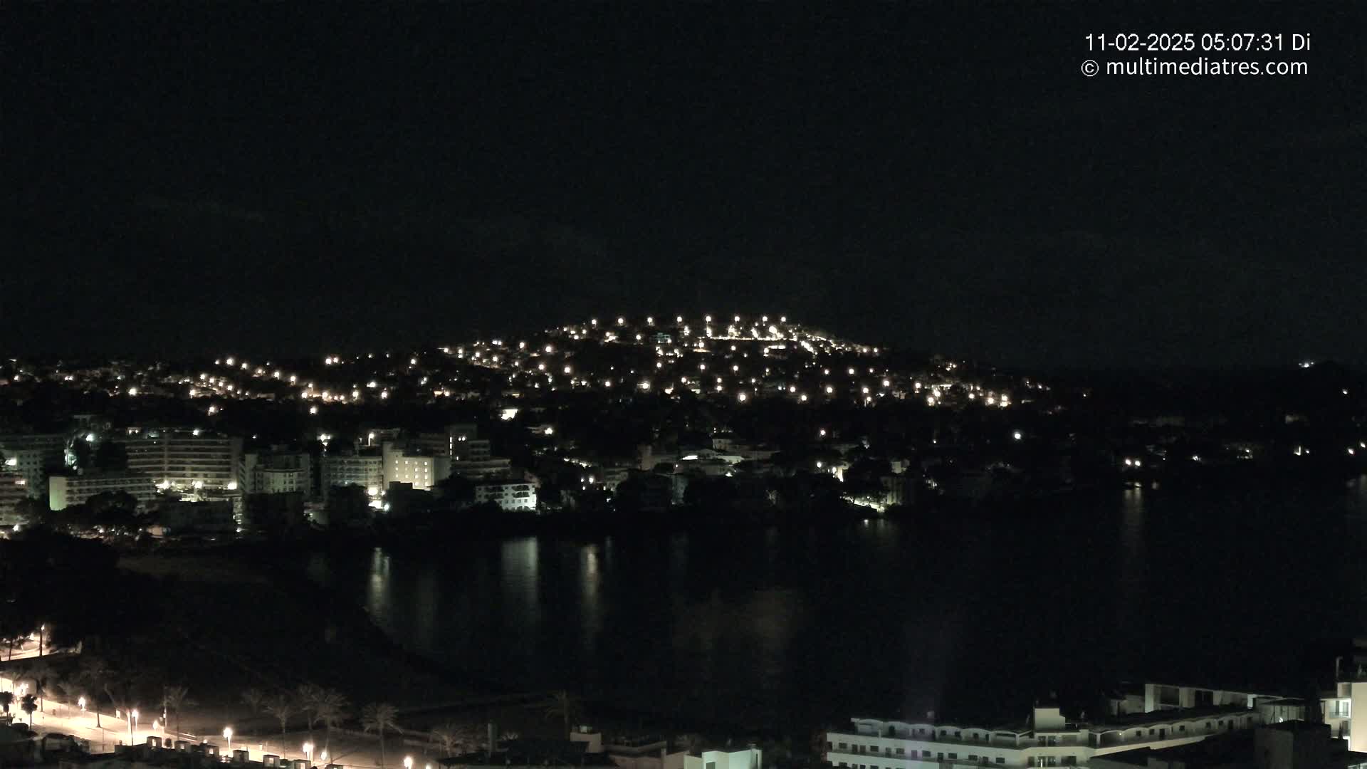 A nighttime view of a city illuminated by lights, situated on a hill overlooking a dark body of water.