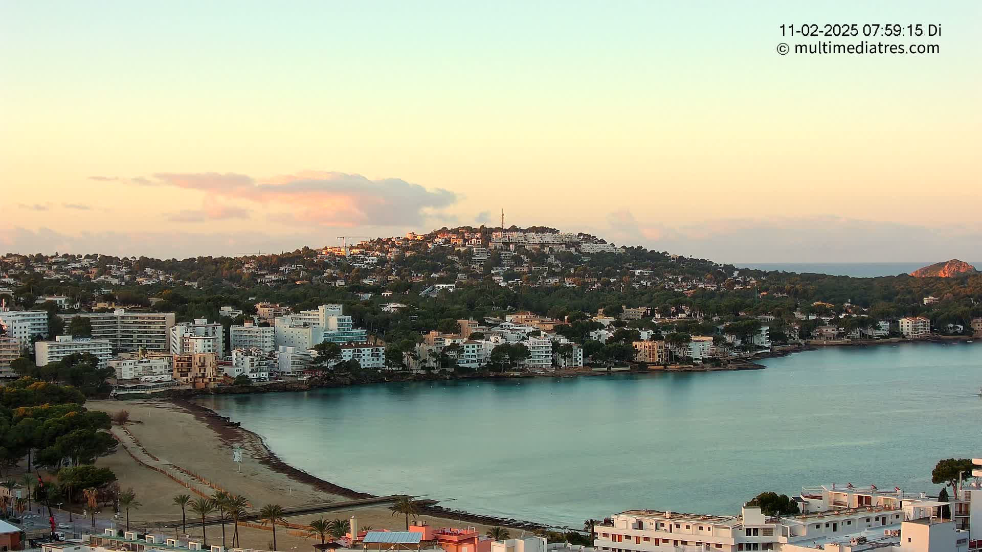 A coastal town at sunrise, nestled around a calm bay with a sandy beach, is situated on a hill covered in greenery.