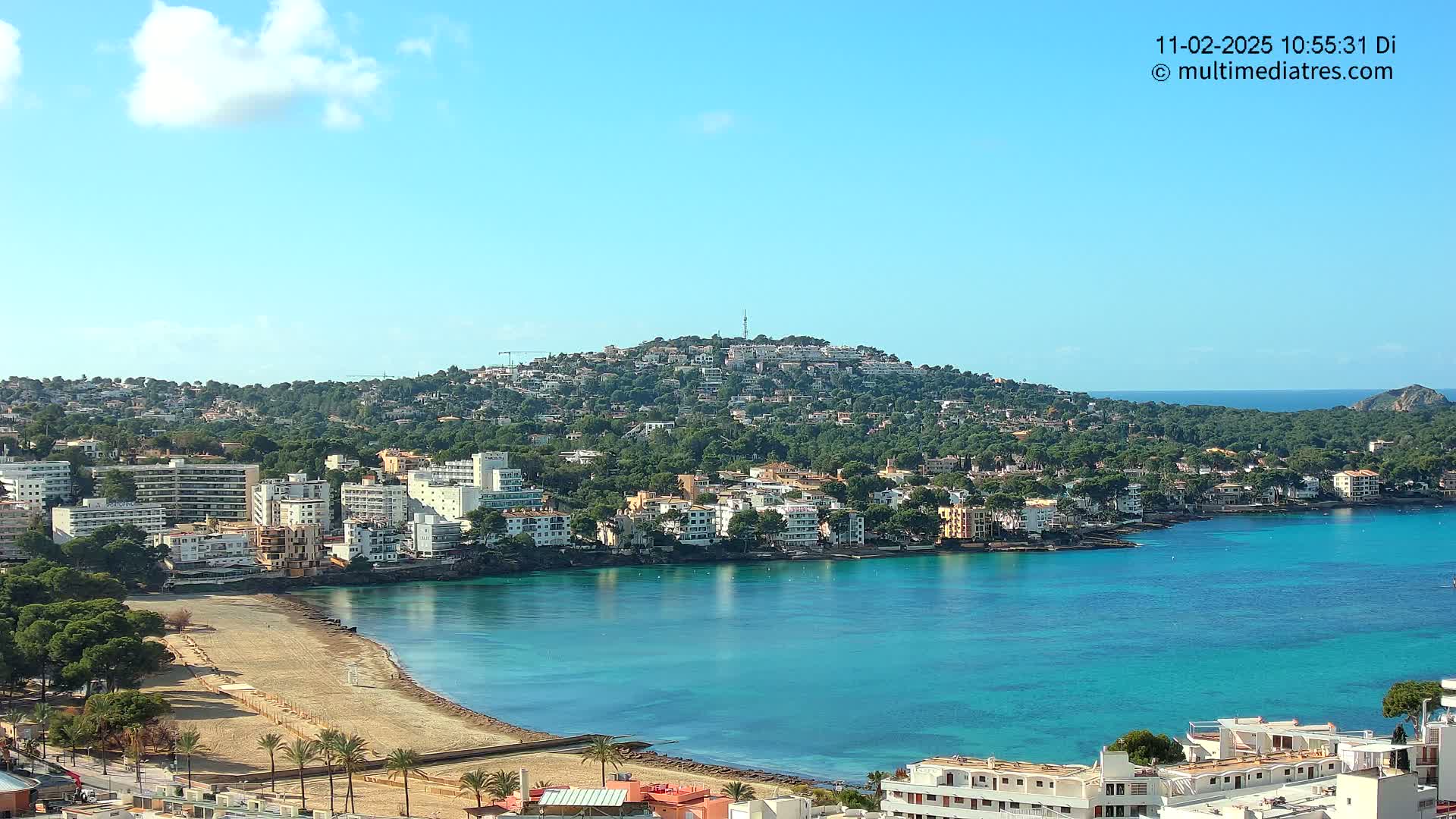 A coastal town with a sandy beach and turquoise water sits below a hill covered in buildings and trees under a mostly sunny sky.