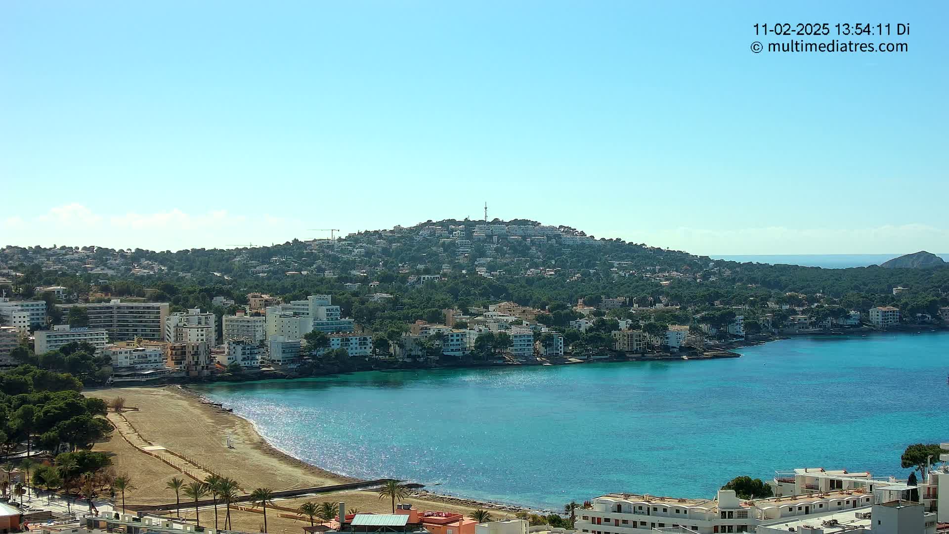 A coastal town nestled against a hill, featuring a sandy beach and clear turquoise water under a bright, sunny sky.