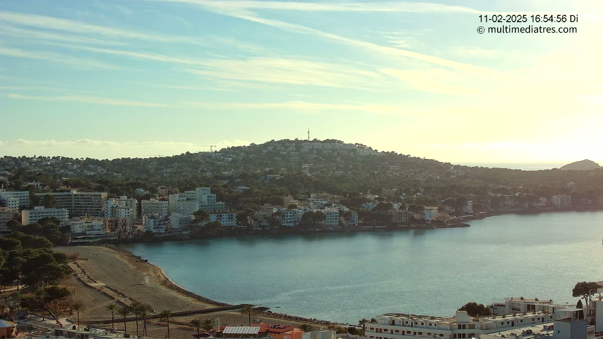 A coastal town nestled against a hill under a mostly clear, light blue sky, with a calm body of water separating the buildings from another hill in the distance.