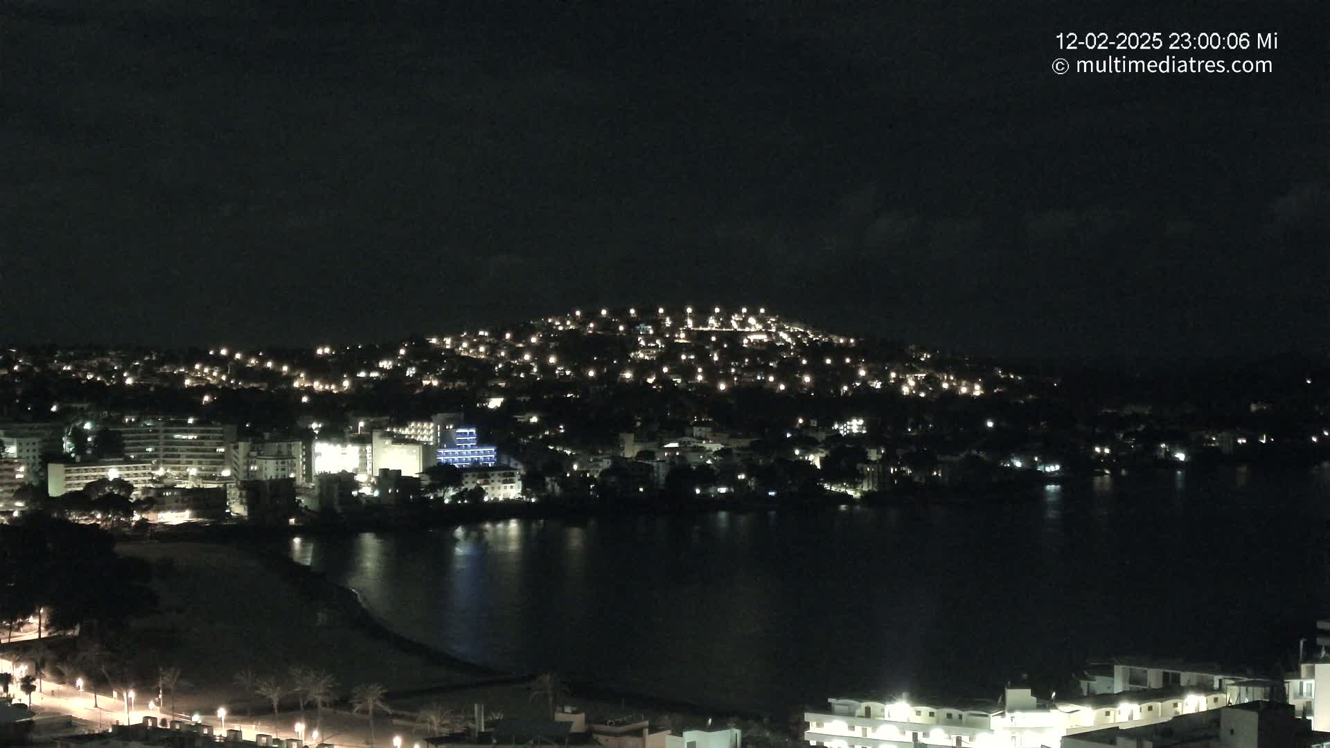 A nighttime view of a city along a dark body of water, illuminated by numerous lights on buildings and streets, under a mostly clear sky.
