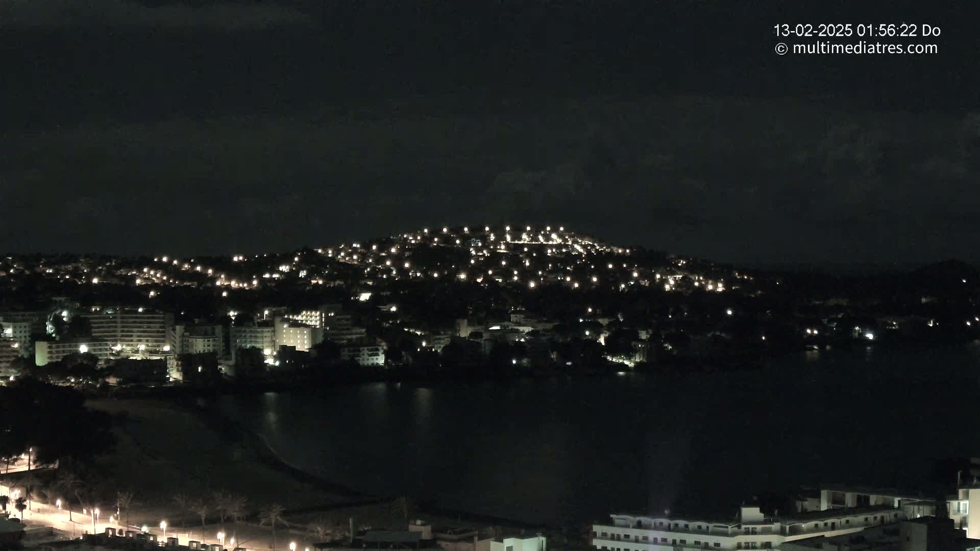 A nighttime view of a city situated on a hill overlooking a dark body of water under a mostly clear sky.