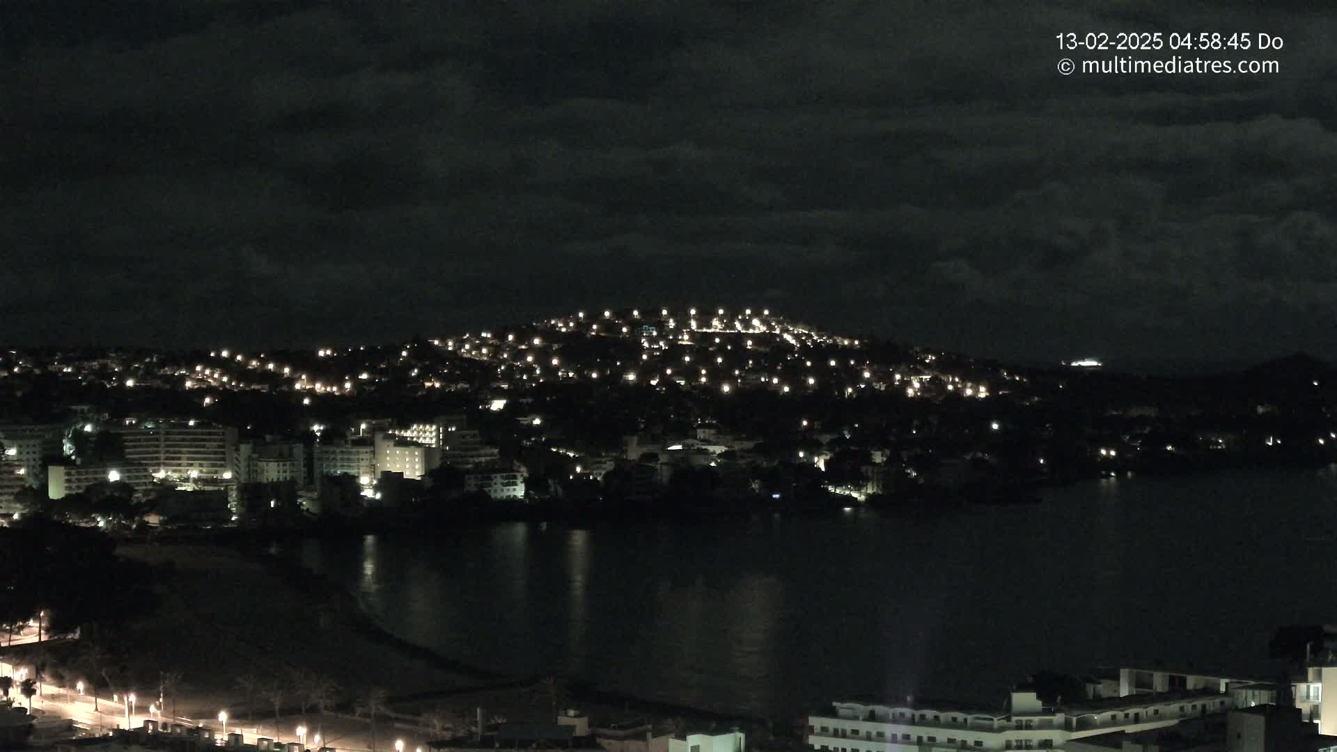 A nighttime view of a coastal town with numerous lights illuminating buildings on a hill overlooking a dark body of water under a cloudy sky.