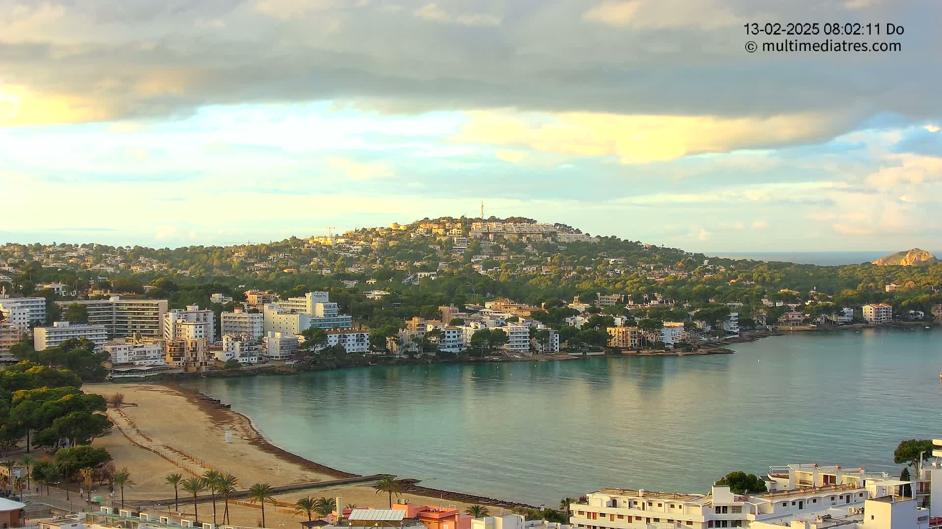 A coastal town nestled around a calm bay, under a partly cloudy sky with hints of sunrise.