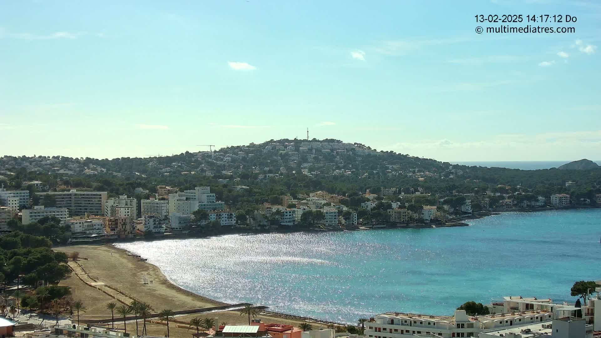 A sunny day reveals a coastal town nestled against a hill, with a calm, sparkling turquoise sea lapping at a sandy beach.