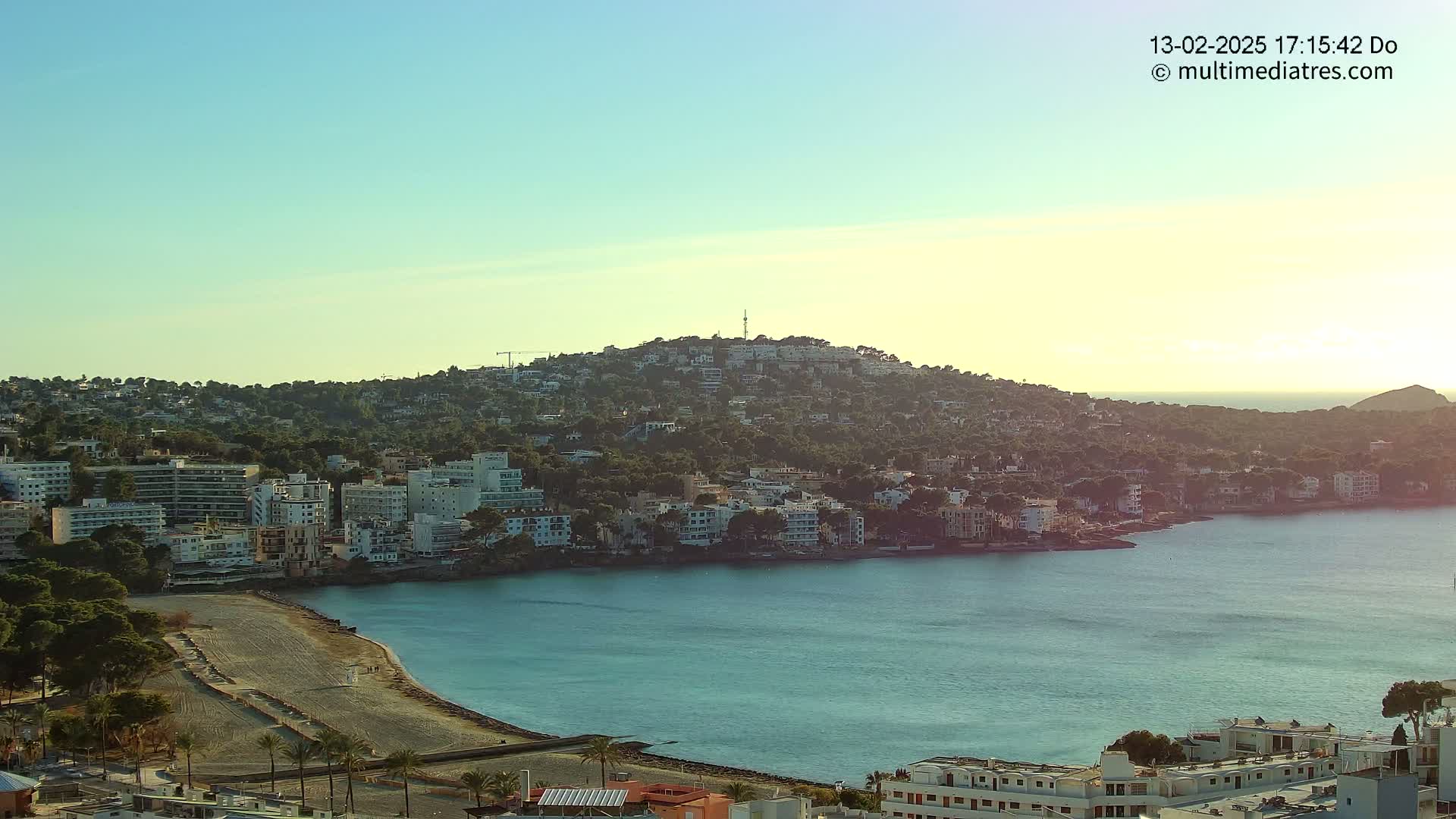 A coastal town nestled against a hill, with calm, teal water and a clear, sunny sky.