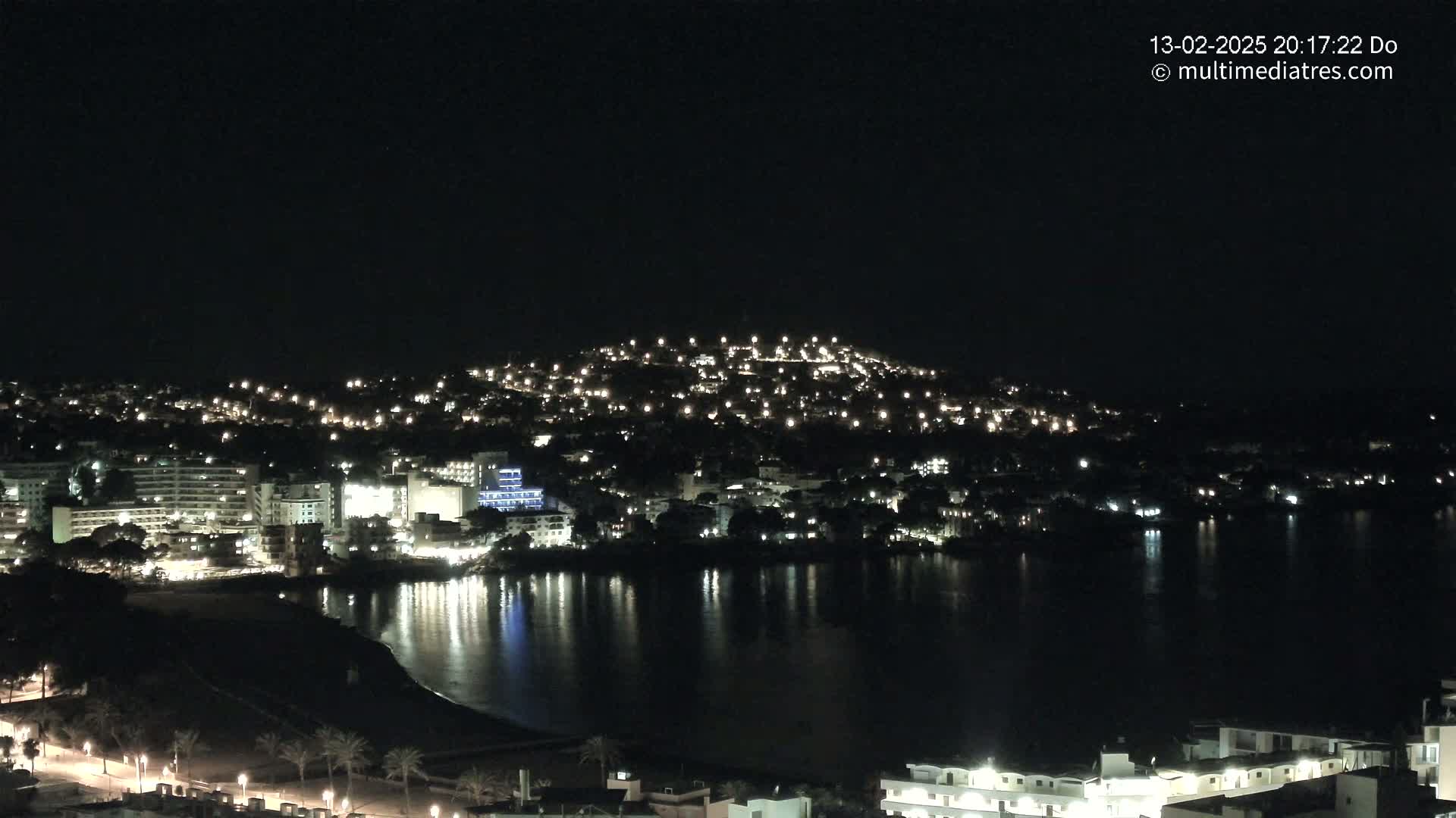 A nighttime view of a coastal town illuminated by lights, reflecting on a calm body of water under a clear, dark sky.