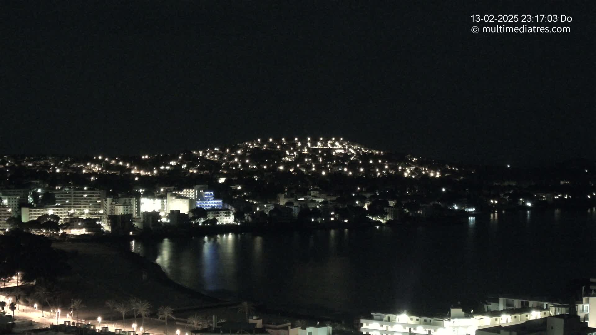 A nighttime view of a coastal town illuminated by many lights, with a dark body of water in the foreground.