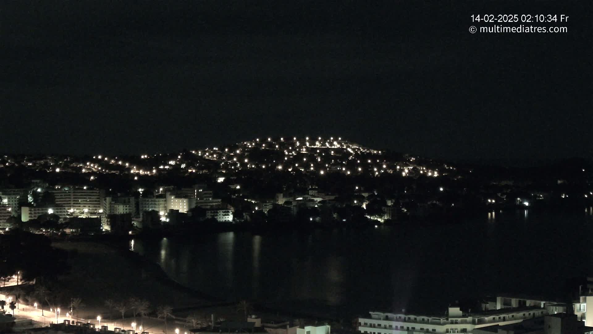 A nighttime view of a city illuminated by lights, situated along a dark body of water, under a clear, dark sky.