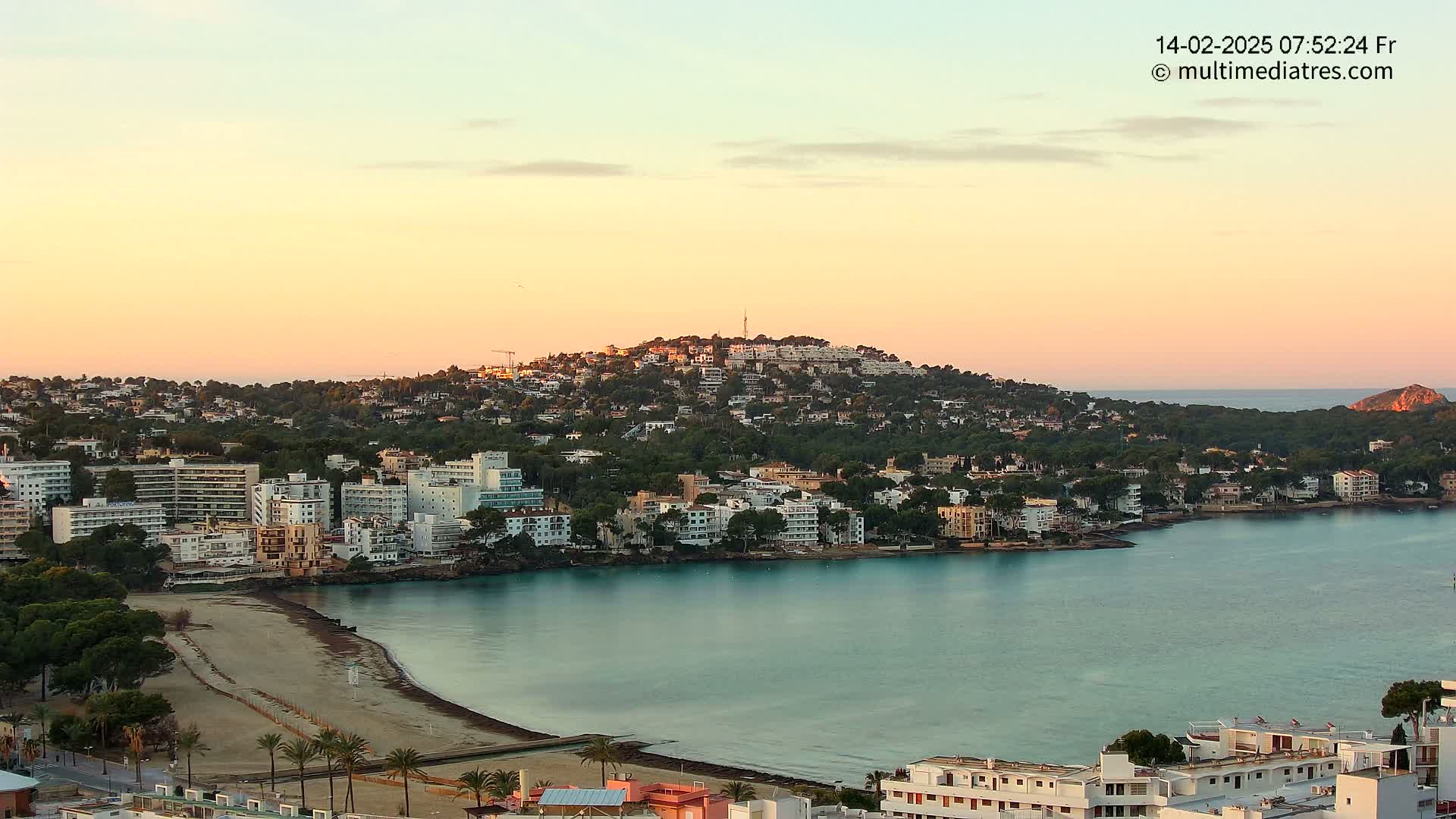 A coastal town at sunrise, nestled around a calm bay with a hill covered in buildings in the background.