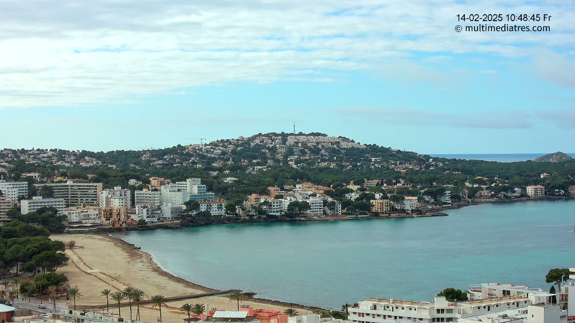 A coastal town with a sandy beach and turquoise water, nestled beneath a hill covered in buildings and trees, under a partly cloudy sky.