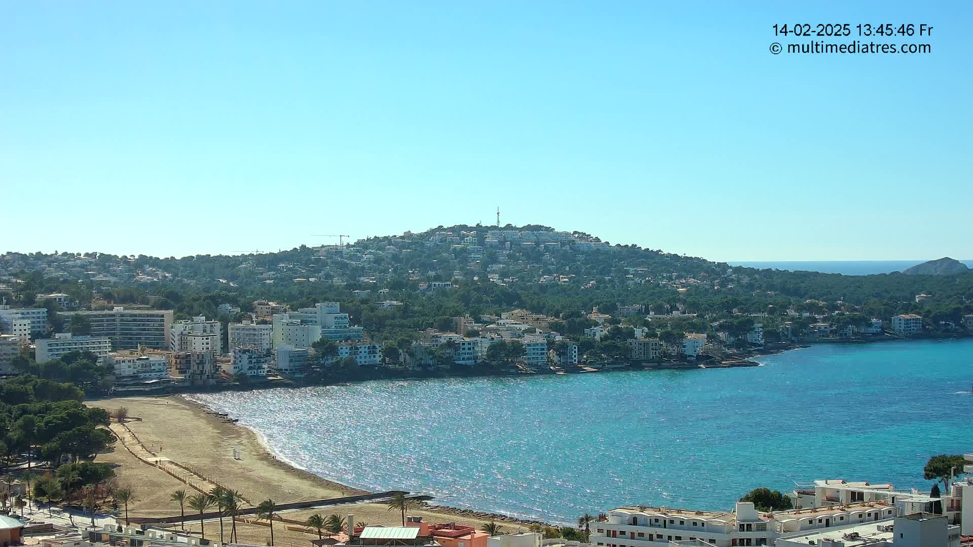 A hilltop town overlooking a calm, sparkling blue bay with a sandy beach.