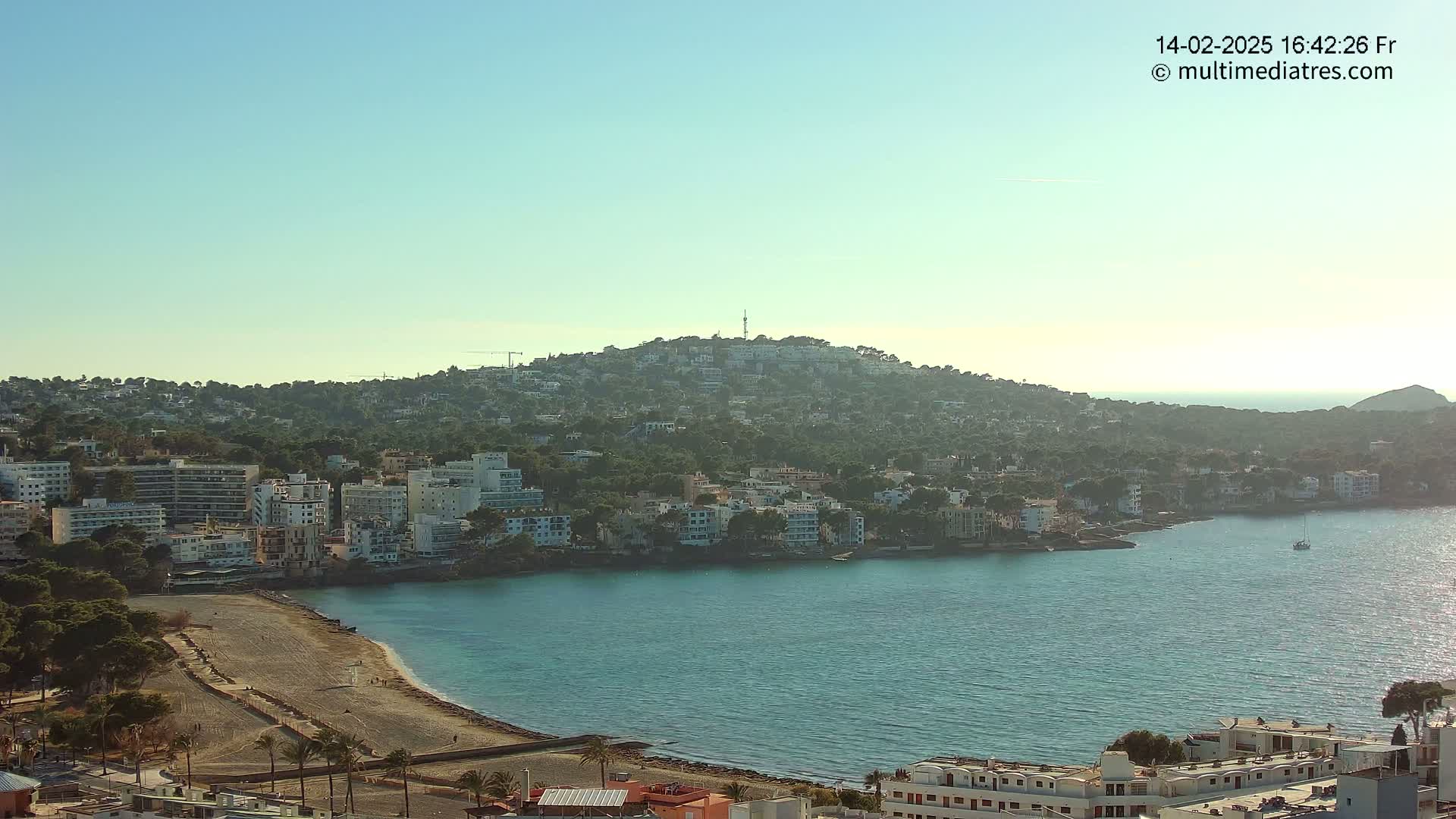 A coastal town nestled against a hill, with a calm, clear body of water in front, under a bright, mostly clear sky.