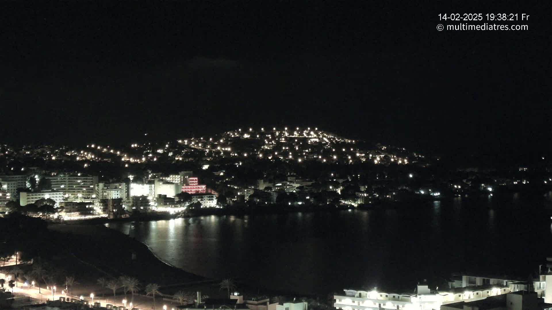 A nighttime coastal town is illuminated, with buildings and lights spread across a hill and along the shoreline, beside a dark body of water.