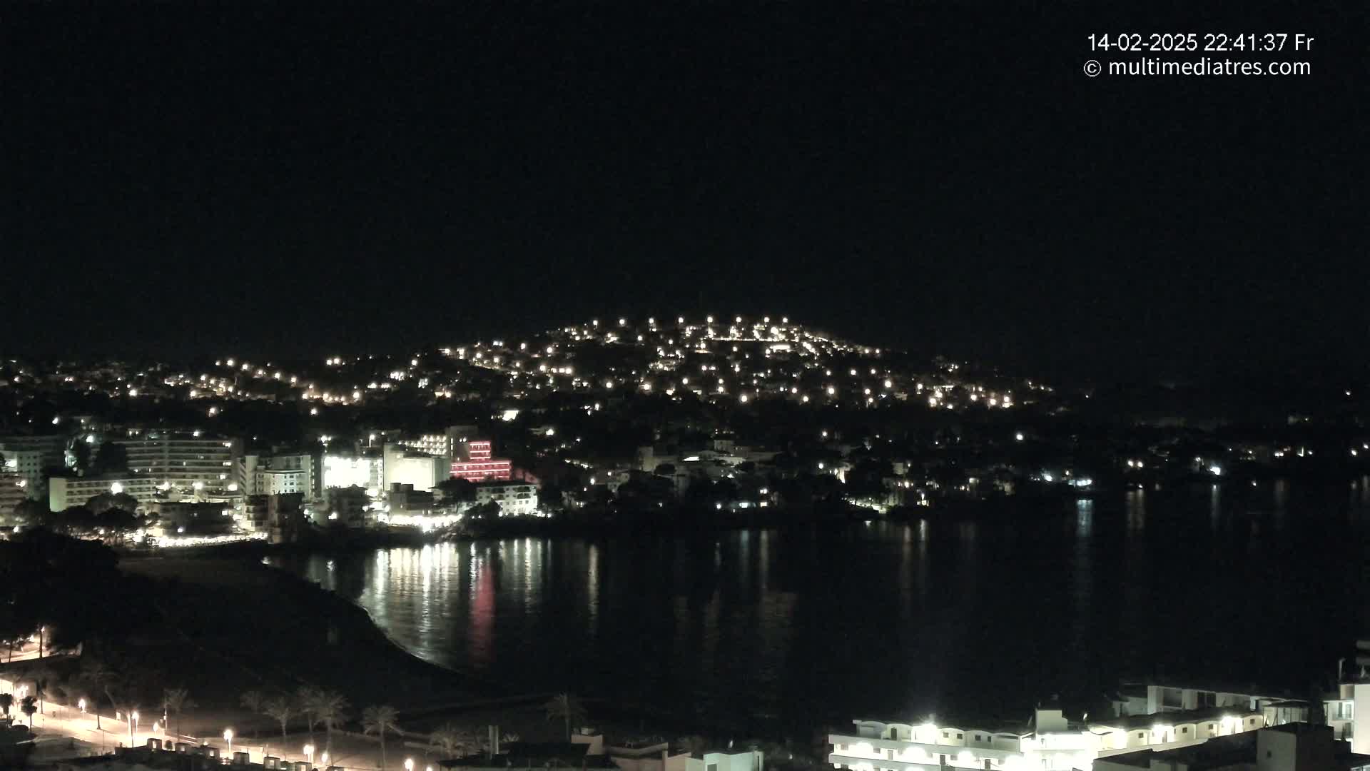 A nighttime view of a coastal town illuminated by lights, with calm water reflecting the city lights.