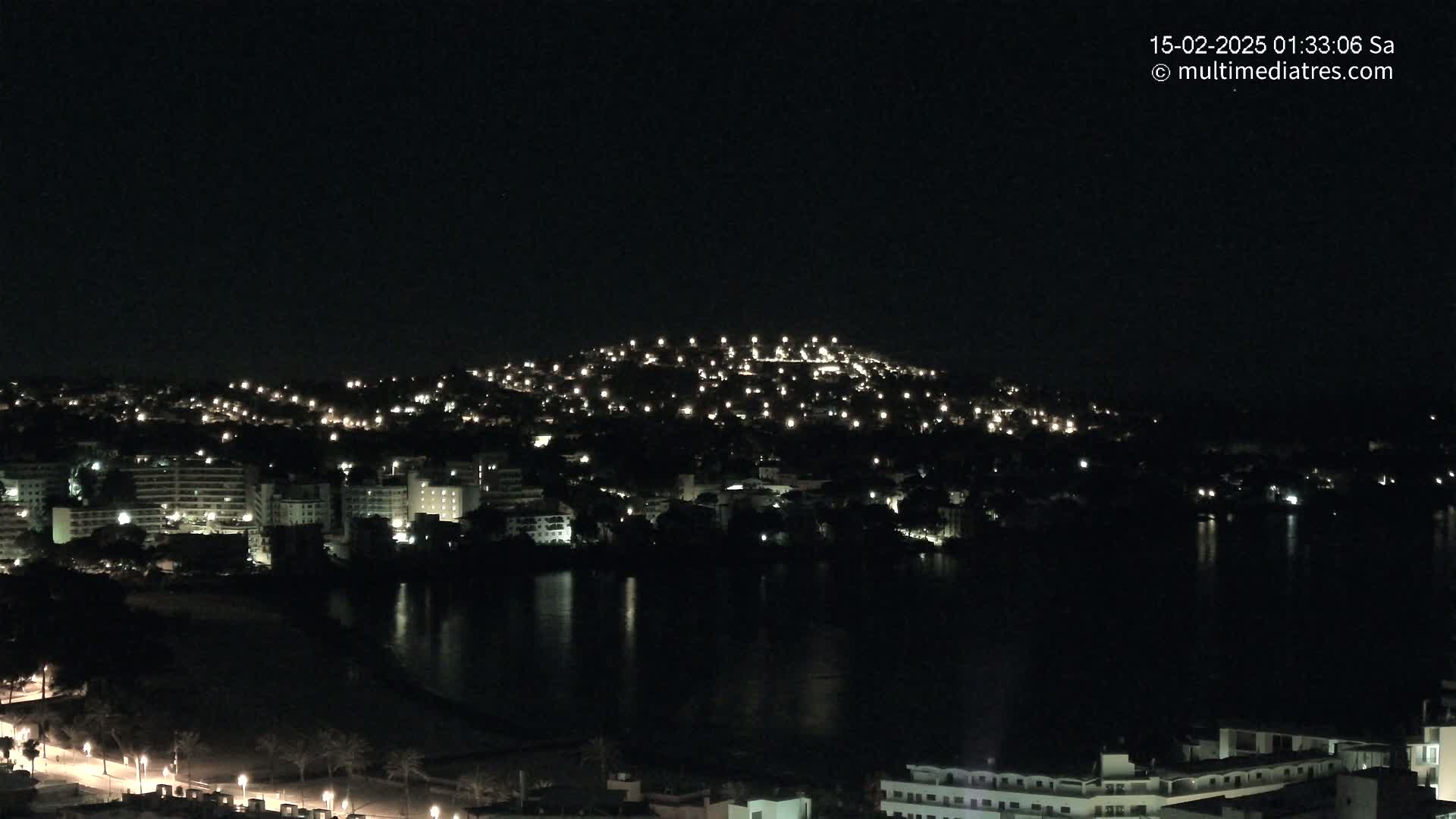 A nighttime view of a city spread across a hill and along a dark body of water, under a clear, starless sky.