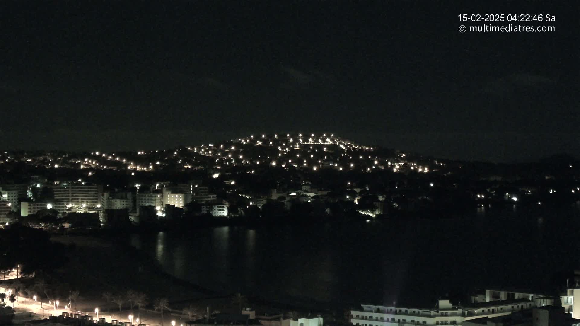 A nighttime view of a city built on a hill and next to a body of dark water, with numerous lights illuminating buildings and streets.