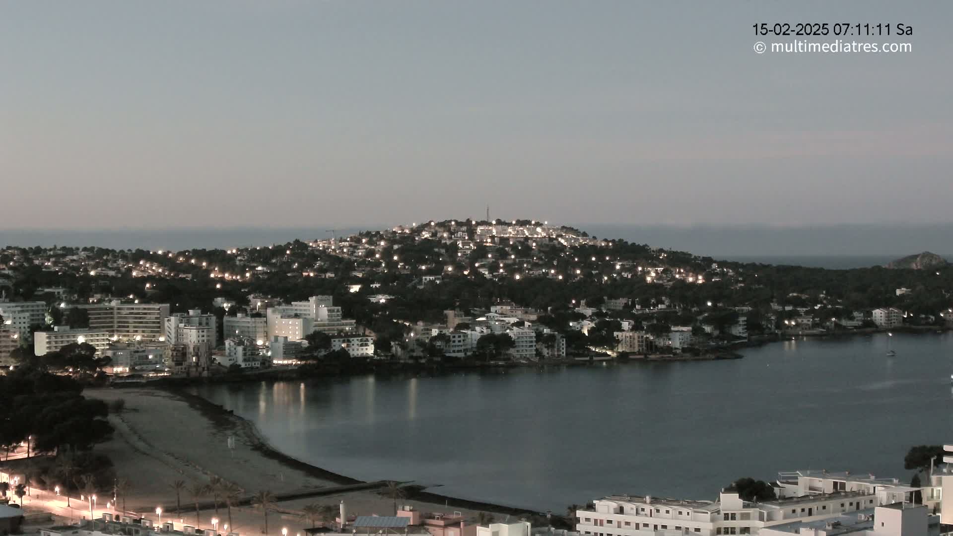 A coastal town with lights illuminated at twilight, nestled on a hill overlooking a calm bay under a dusky sky.