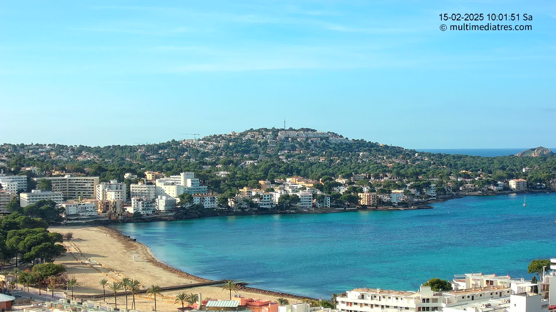 A coastal town nestled against a verdant hill overlooking a calm, turquoise bay under a clear blue sky.