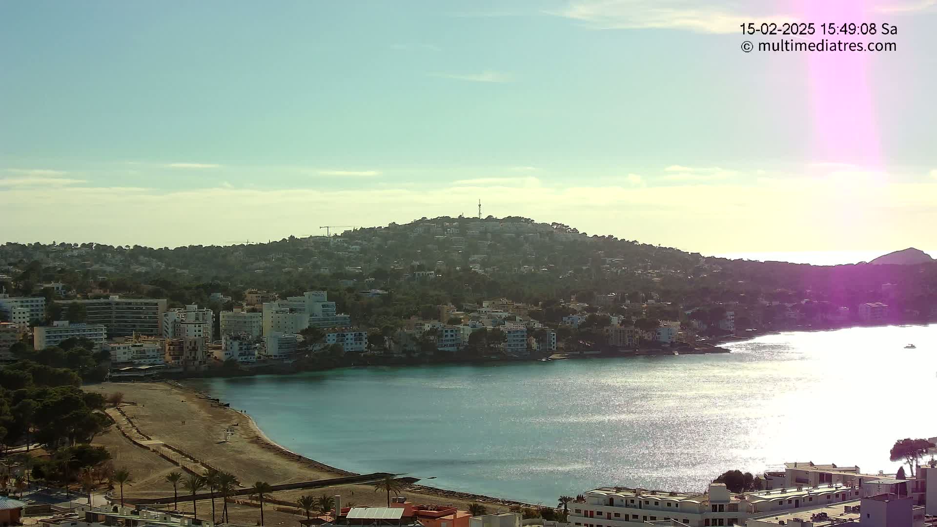 A coastal town nestled on a hillside with a calm, sunlit bay under a mostly clear sky.