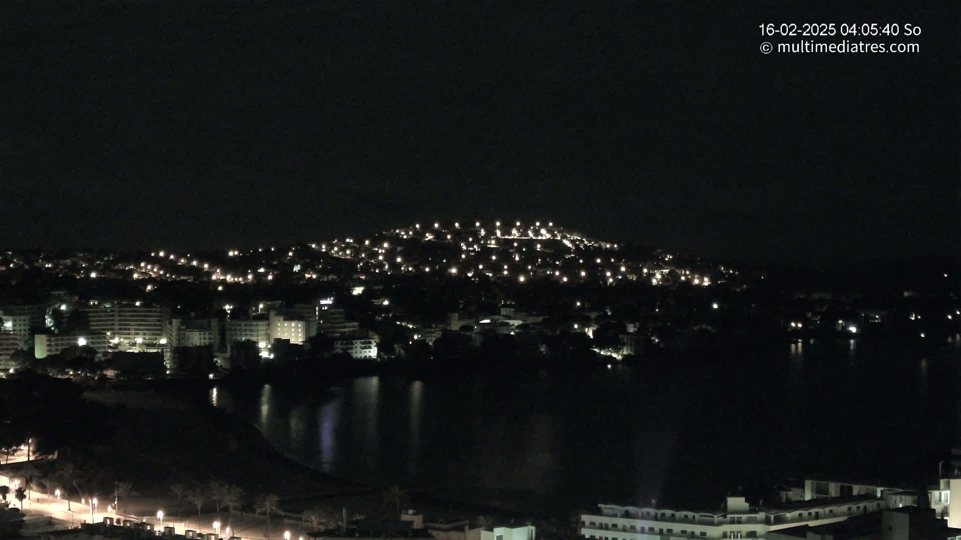 A nighttime view of a coastal town illuminated by numerous lights, with a dark body of water reflecting the lights in the foreground.