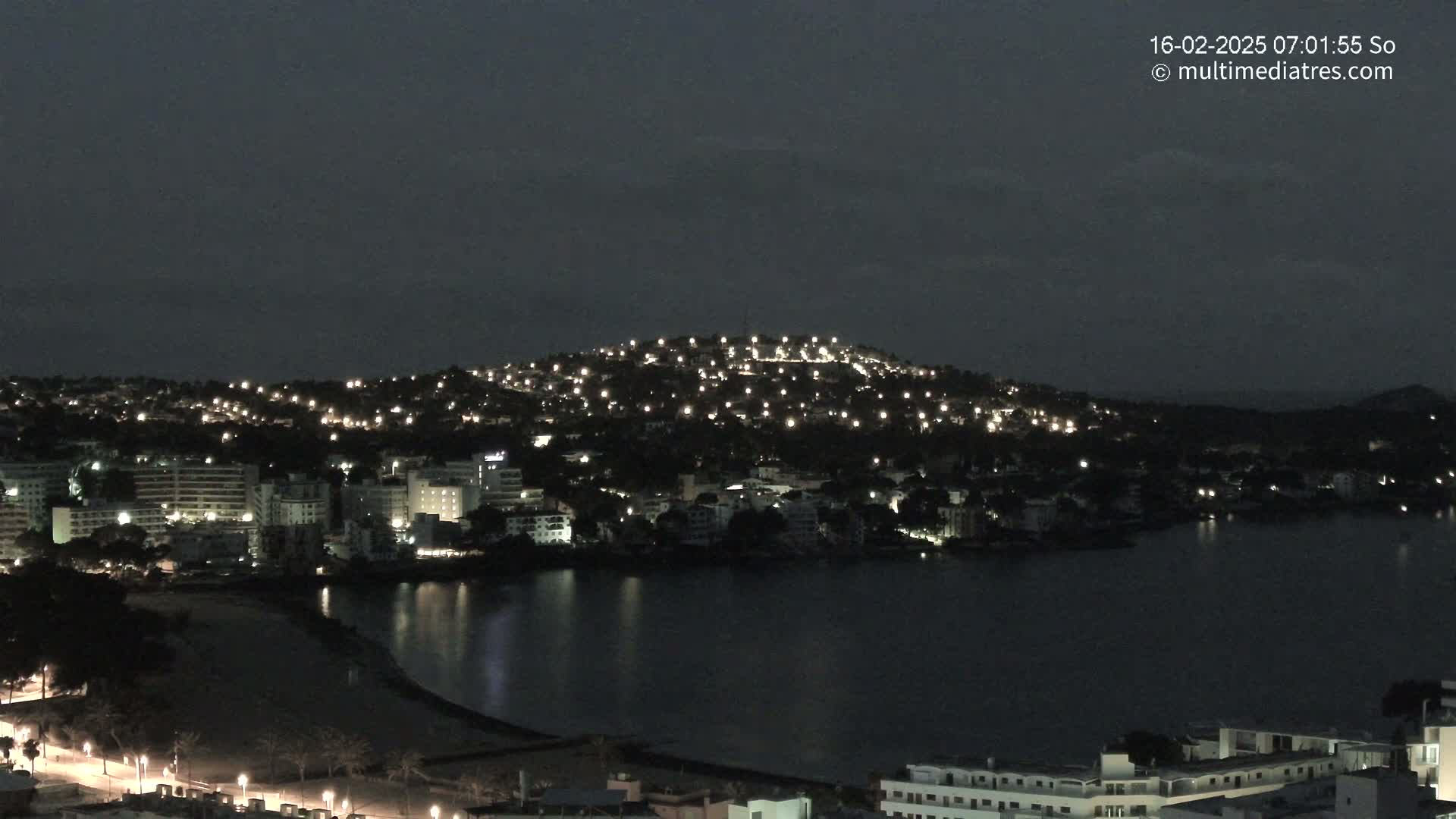 A nighttime view of a coastal town illuminated by numerous lights, nestled against a dark hill and bordering a calm, dark body of water under a mostly clear, dark sky.