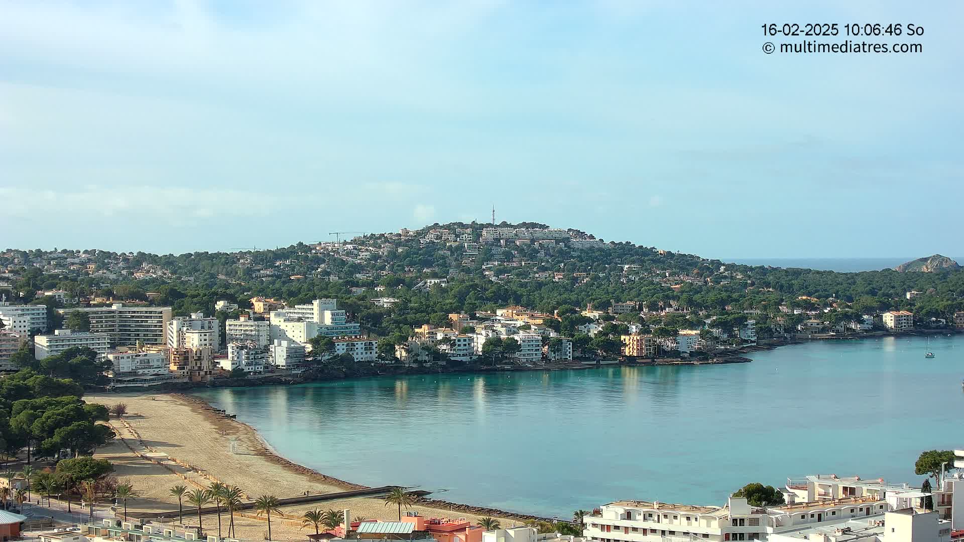 A coastal town nestled against a verdant hill, with a calm bay of turquoise water and a sandy beach, under a mostly clear sky.