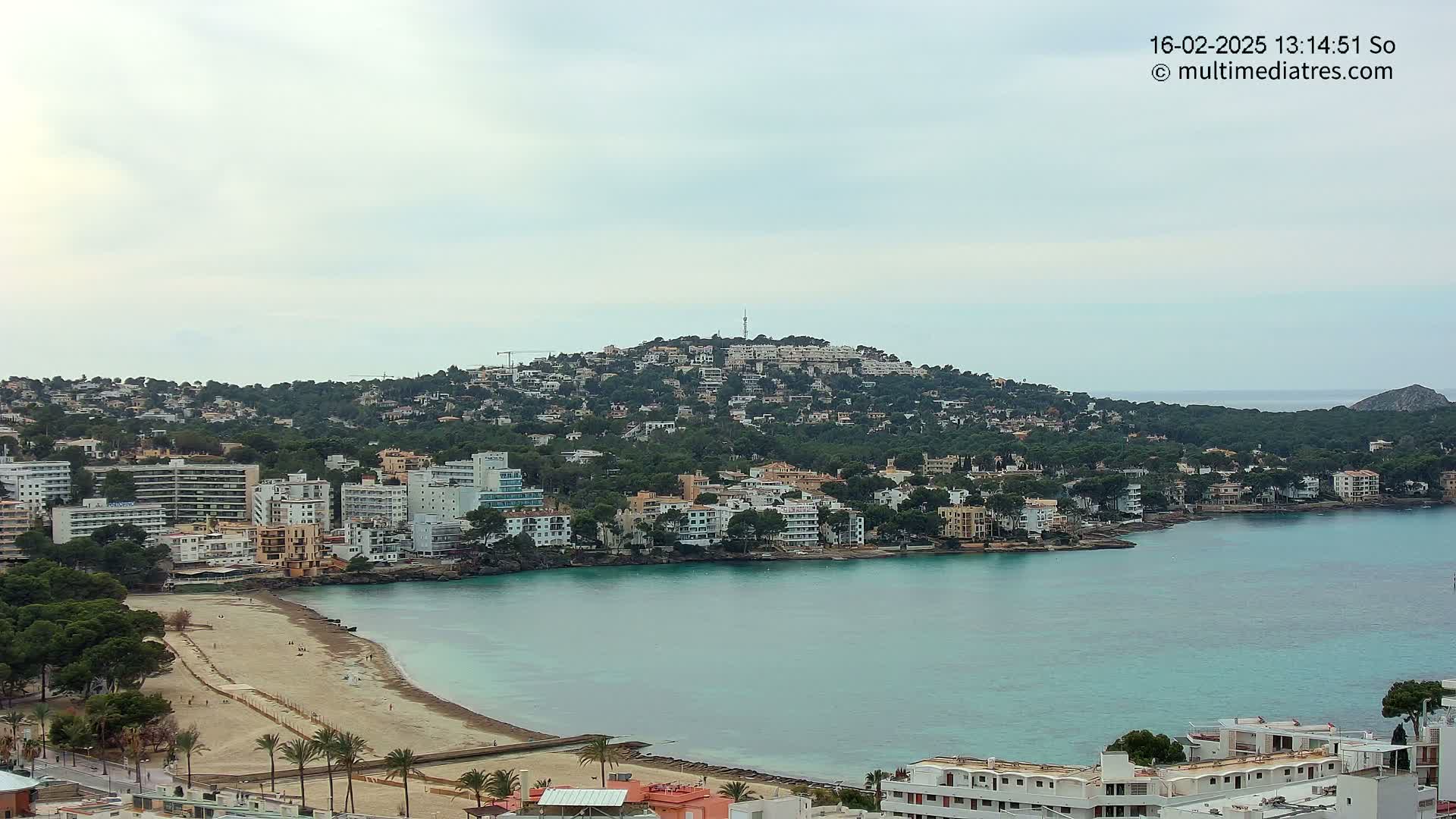 A coastal town with white buildings sprawls across a hill and along a sandy beach bordering a calm, turquoise bay under an overcast sky.