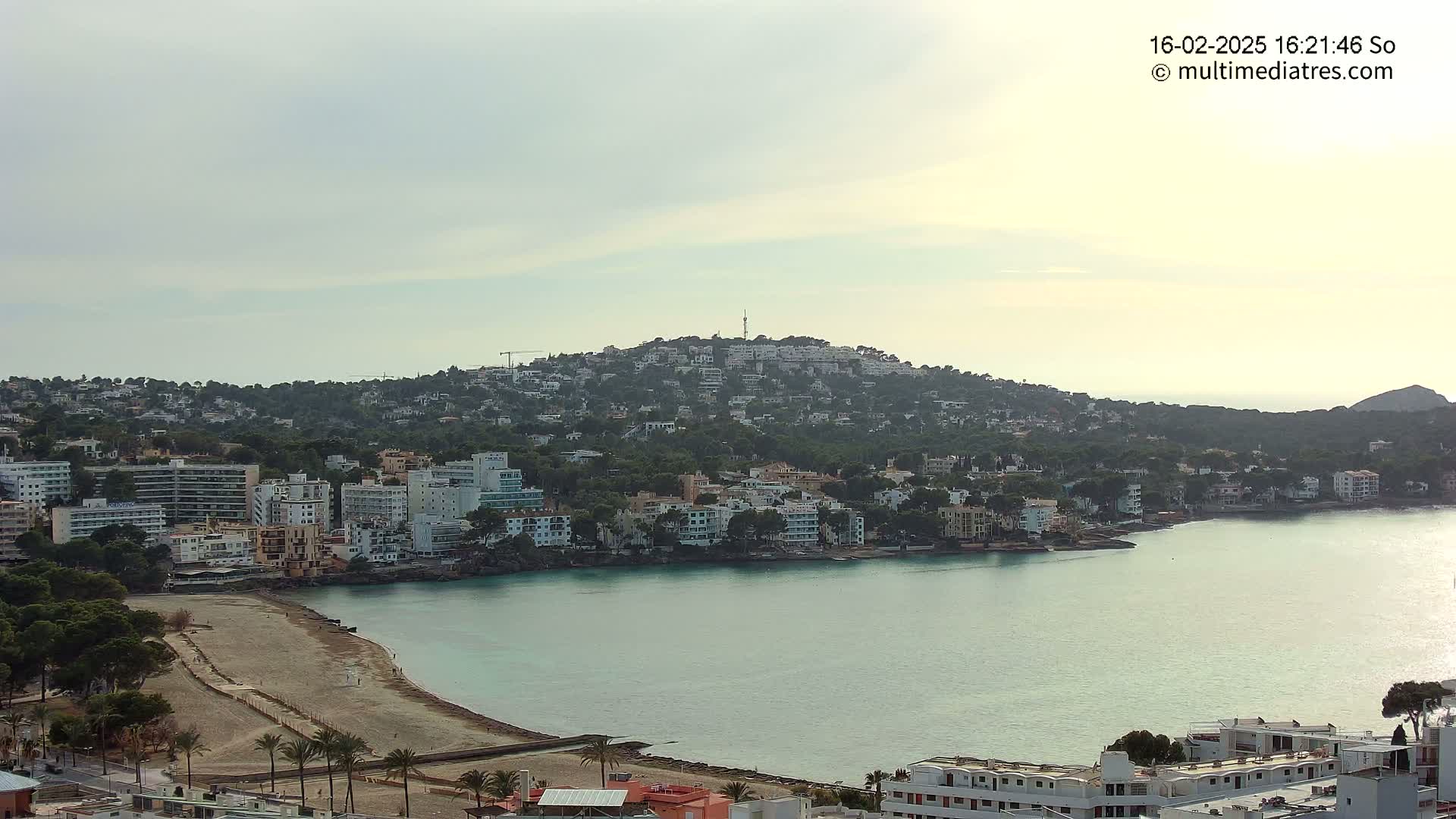 A coastal town nestled on a hill overlooking a calm, teal bay under a cloudy sky.