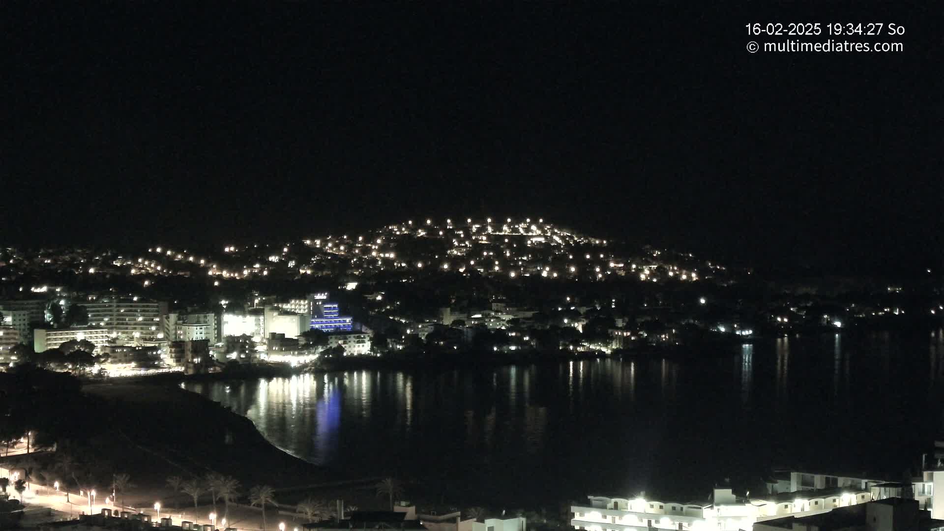 A nighttime view of a town nestled on a hillside overlooking a calm body of water, with numerous lights illuminating buildings and streets.