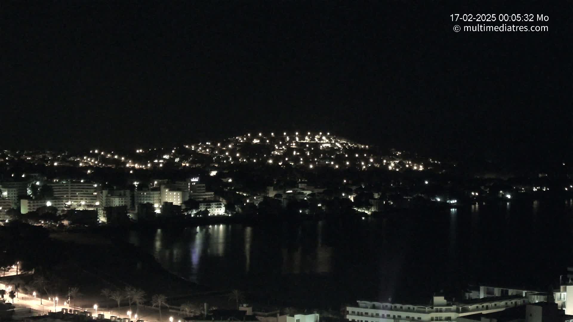 A nighttime view of a city illuminated by lights, situated along a dark body of water under a clear, dark sky.