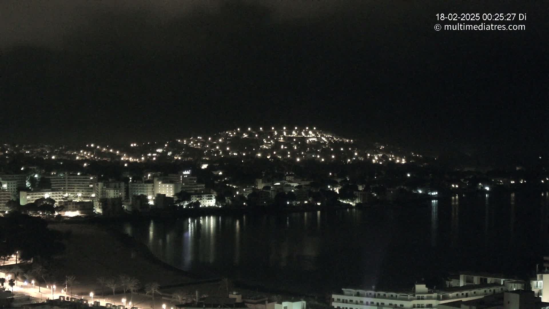 A nighttime view of a coastal town with numerous lights illuminating buildings and hillsides, reflected in dark water under a cloudy sky.