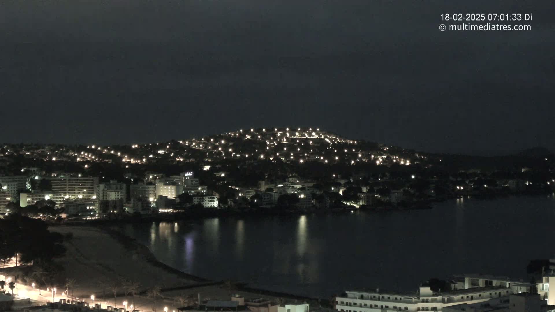 A nighttime view of a city on a hill overlooking a dark body of water under a cloudy sky.