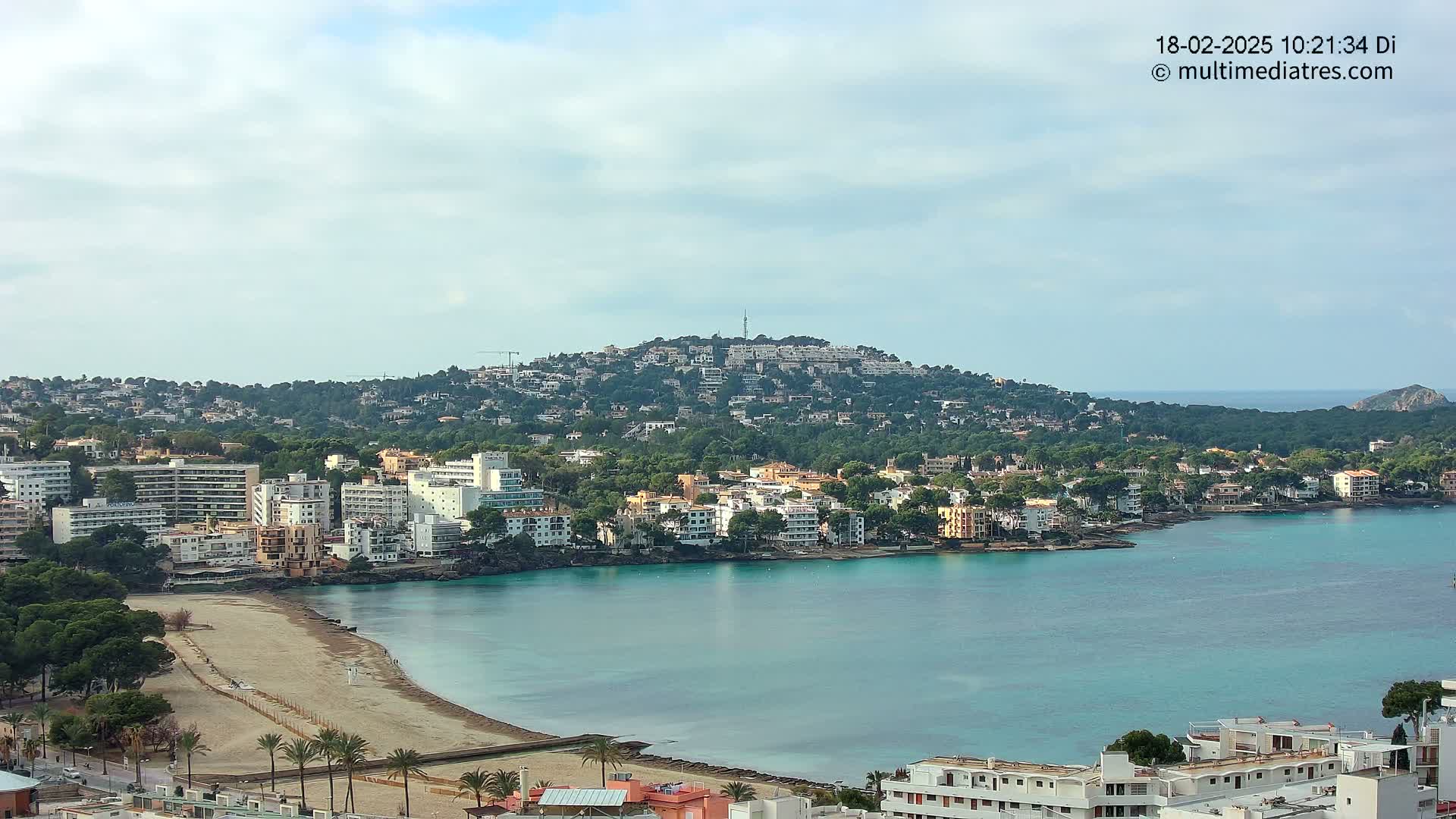 A coastal town with a sandy beach and turquoise water, nestled beneath a hill covered in buildings and greenery, under a mostly cloudy sky.