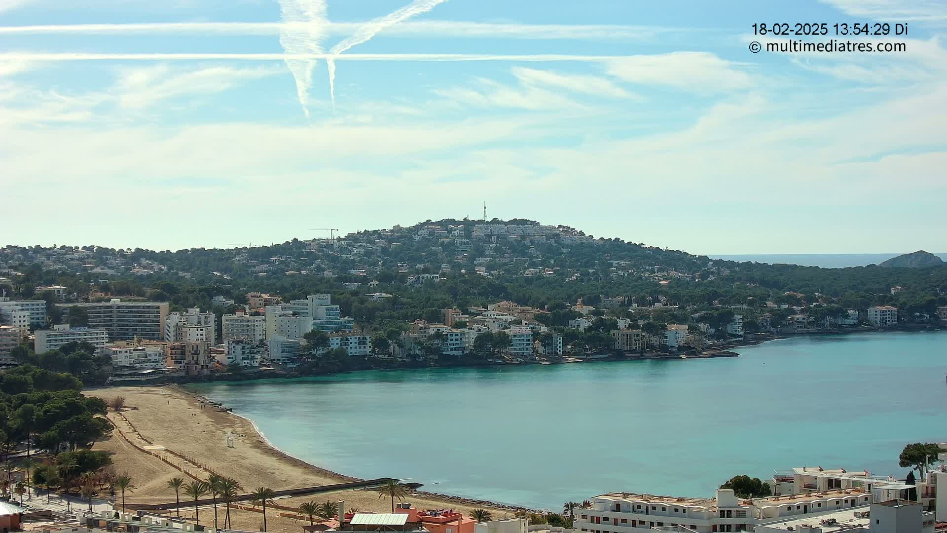 A coastal town with a sandy beach and turquoise water under a mostly sunny sky with contrails.