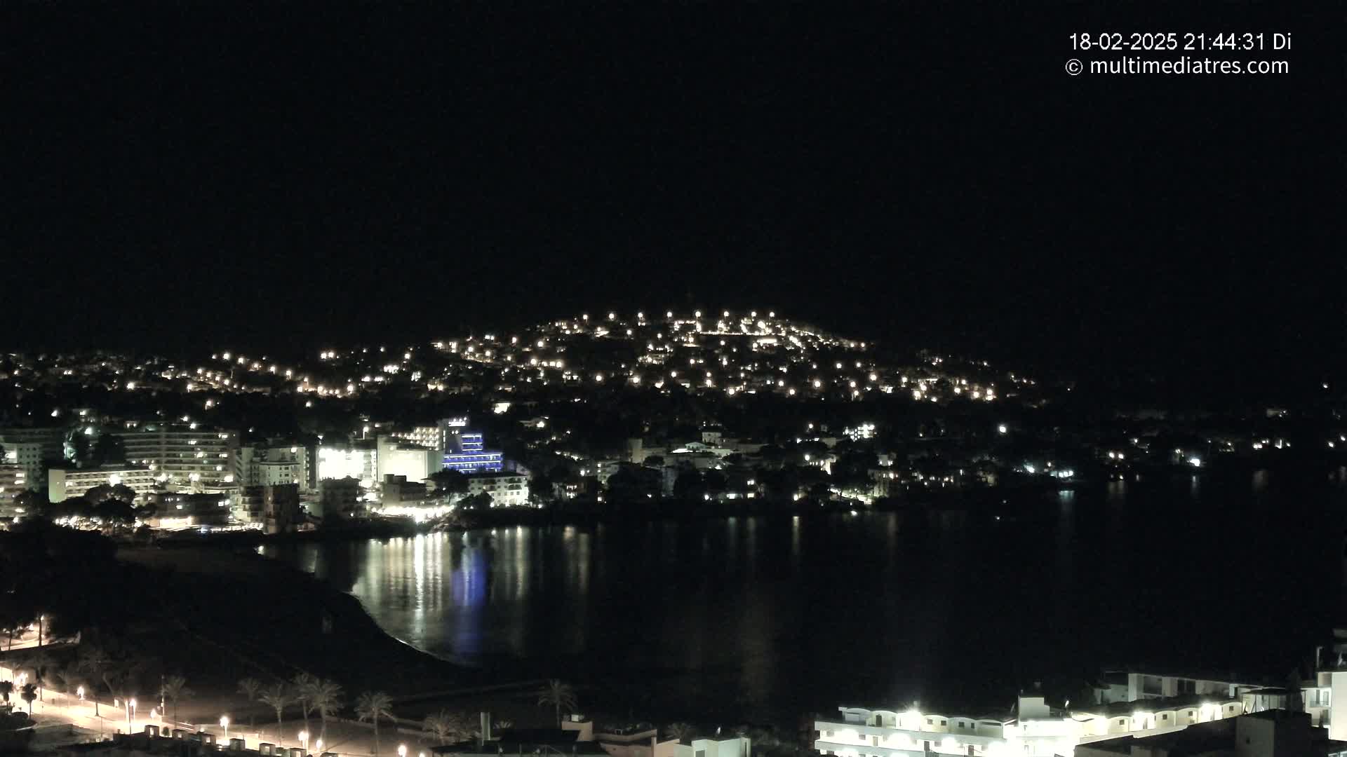 A nighttime view of a coastal town illuminated by numerous lights, reflecting on a dark body of water under a clear, dark sky.