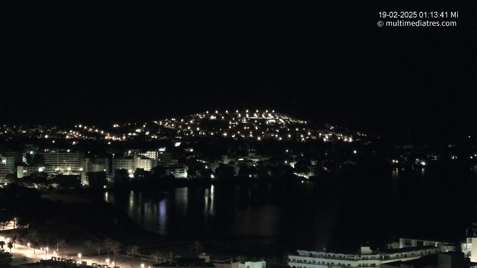 A nighttime view of a coastal town illuminated by many lights, reflecting in a dark body of water under a clear, dark sky.