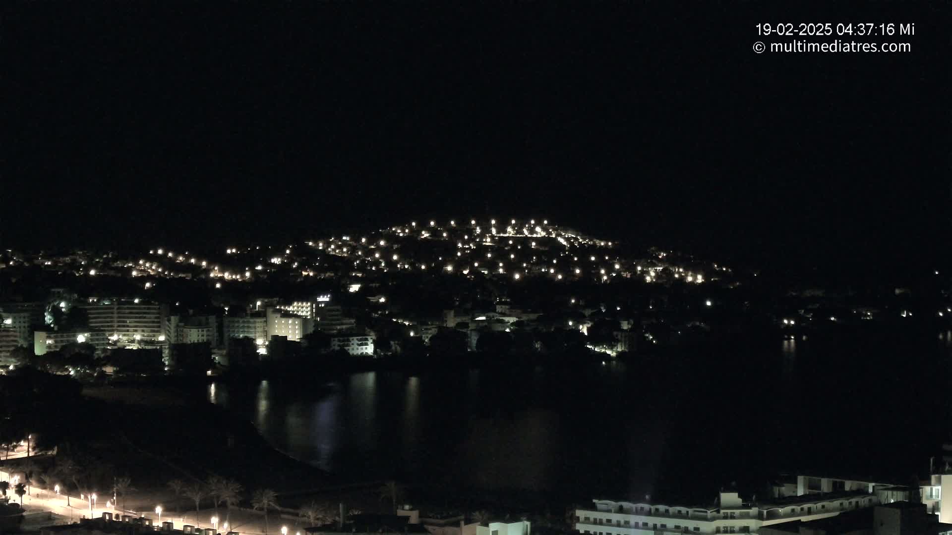 A nighttime view of a city illuminated by numerous lights, situated beside a dark body of water under a clear, dark sky.
