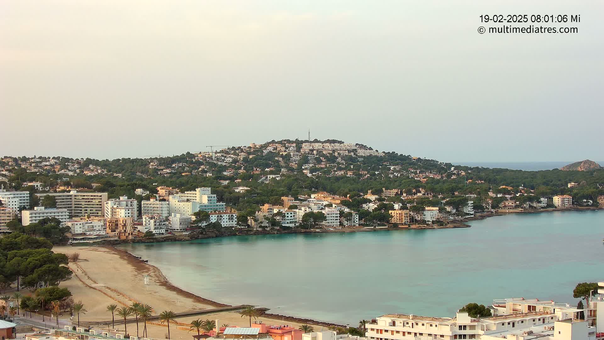 A coastal town with buildings nestled on a hillside overlooking a calm, turquoise bay with a sandy beach.