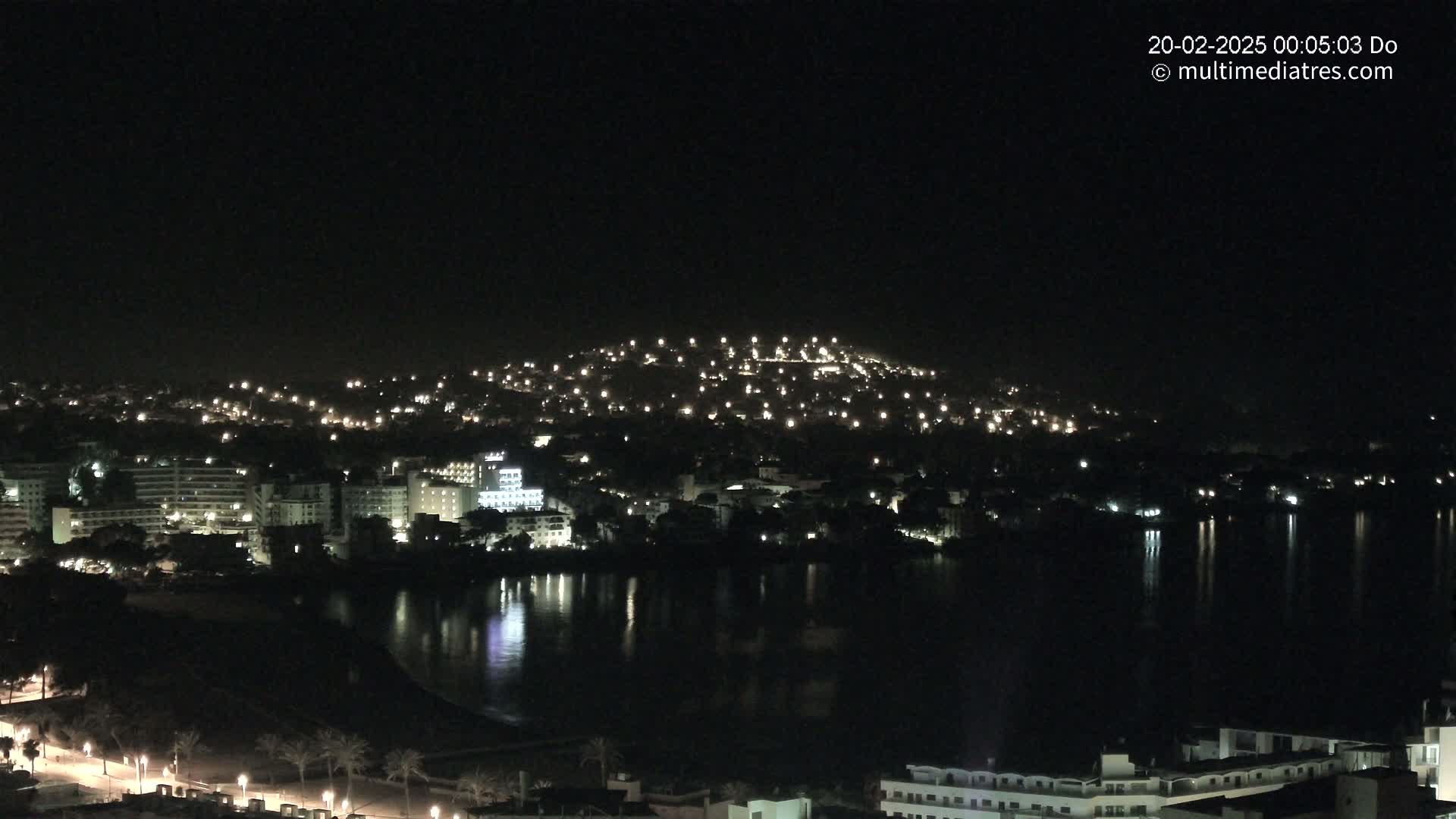 A nighttime view of a coastal town with many lights reflecting on a calm body of water.