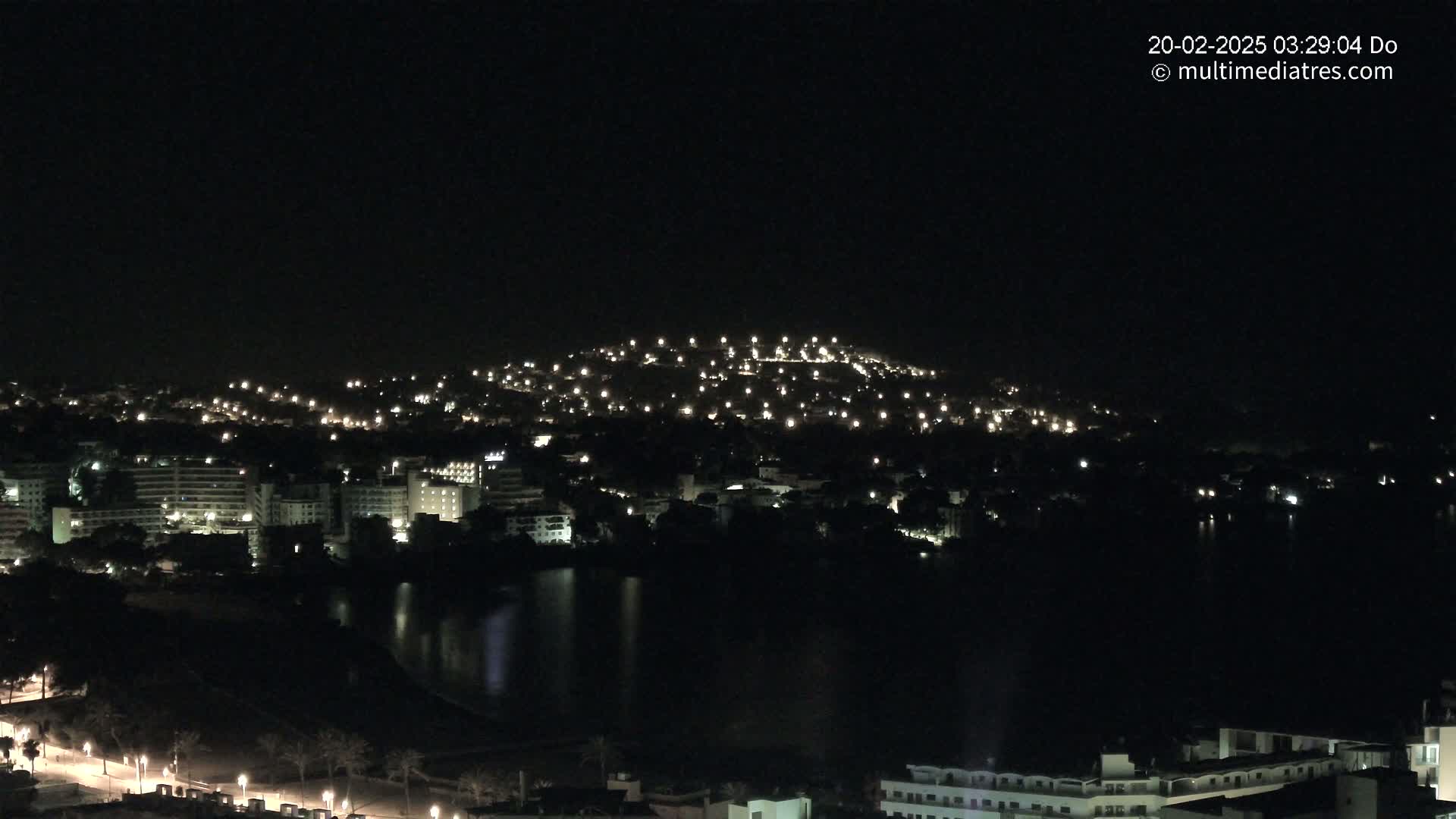A nighttime view of a city nestled beside a dark body of water, with numerous lights illuminating buildings and a hill in the background.