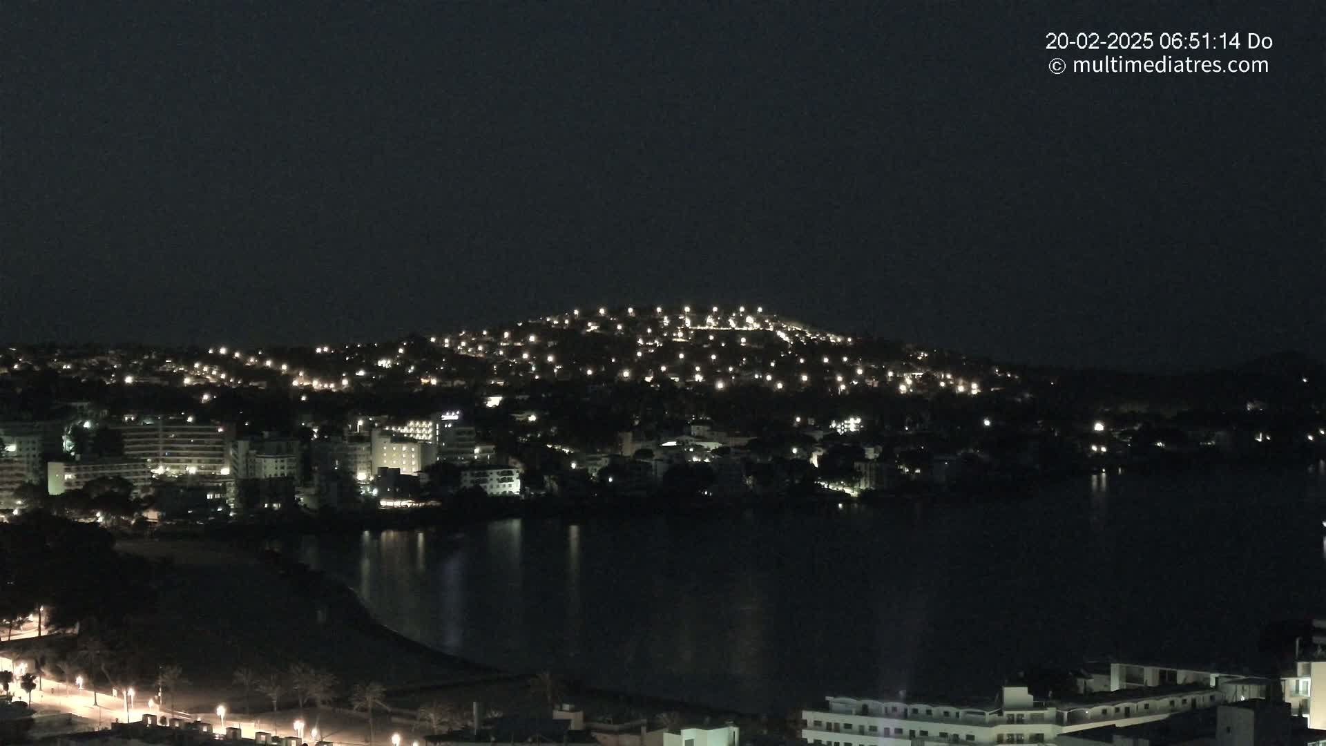 A nighttime view shows a city of buildings illuminated by lights along a dark body of water, with a hill dotted with lights in the background under a clear, dark sky.