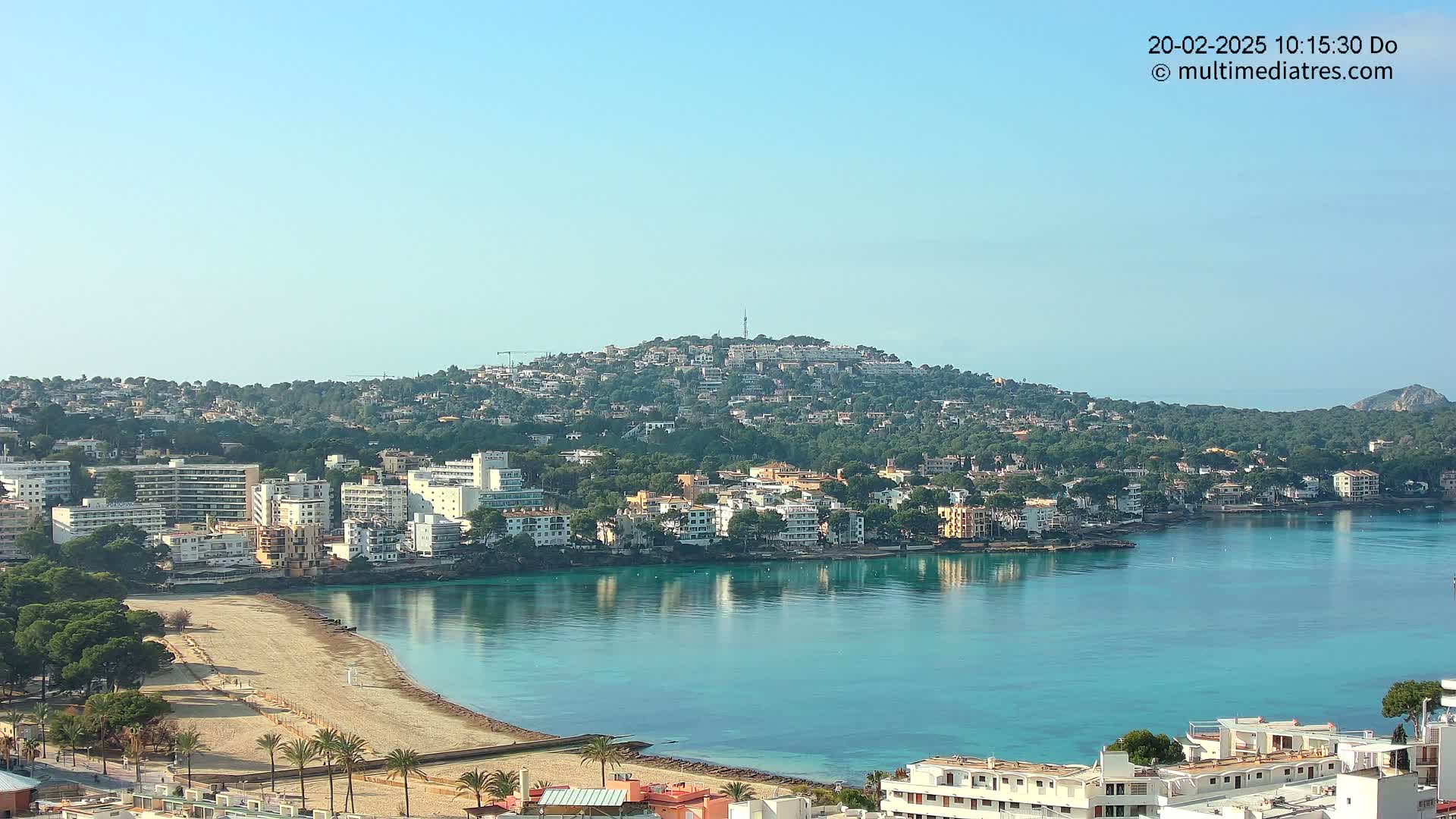 A clear, sunny day reveals a coastal town nestled against a hillside, with a calm, turquoise bay stretching out to a sandy beach.