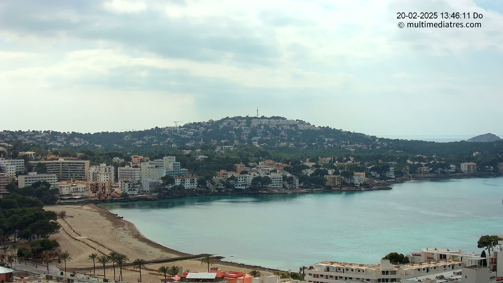 A coastal town nestled against a hill, with a sandy beach and calm, turquoise water under a mostly cloudy sky.