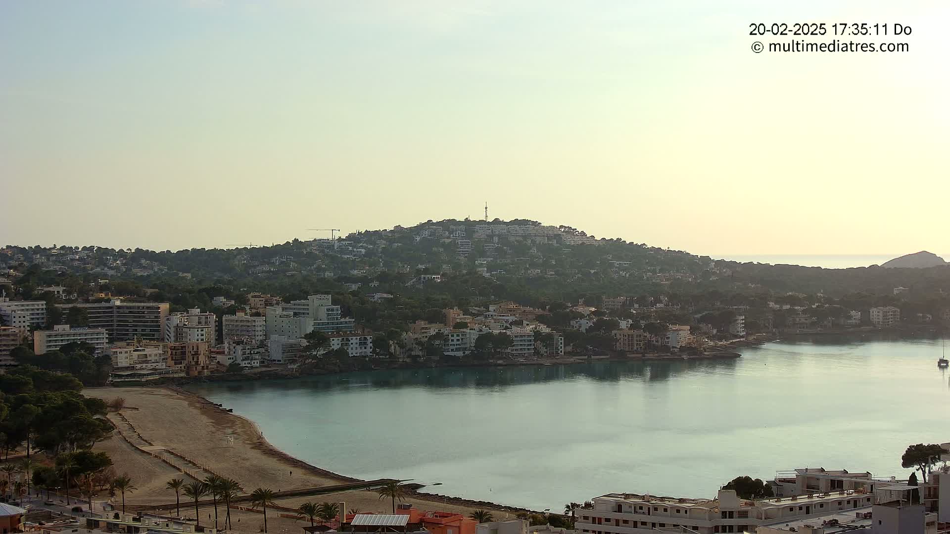 A coastal town nestled on a hillside overlooking a calm bay under a clear, light sky.