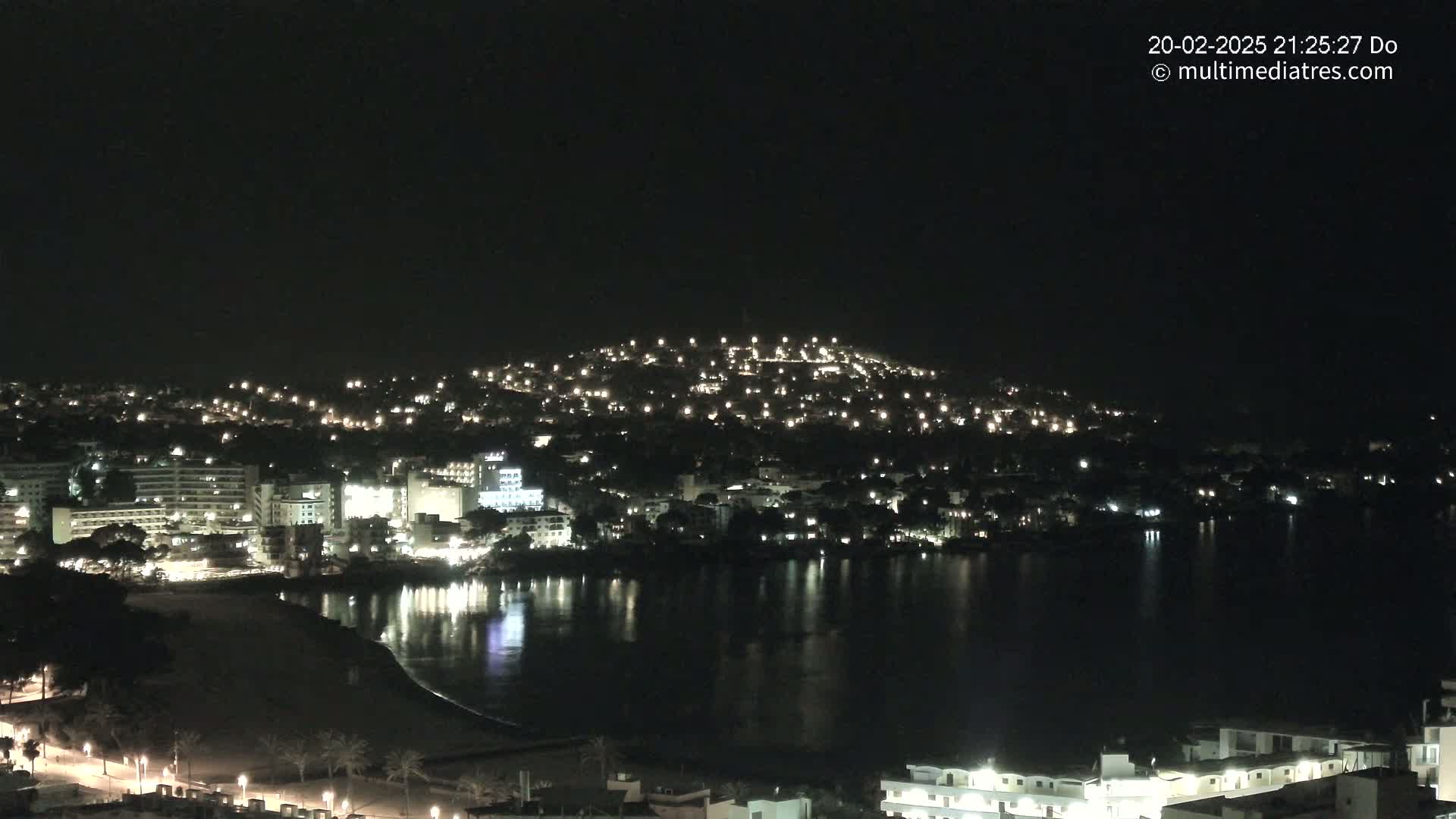 A nighttime view of a coastal town illuminated by lights, nestled against a hill, with a calm body of water reflecting the lights.