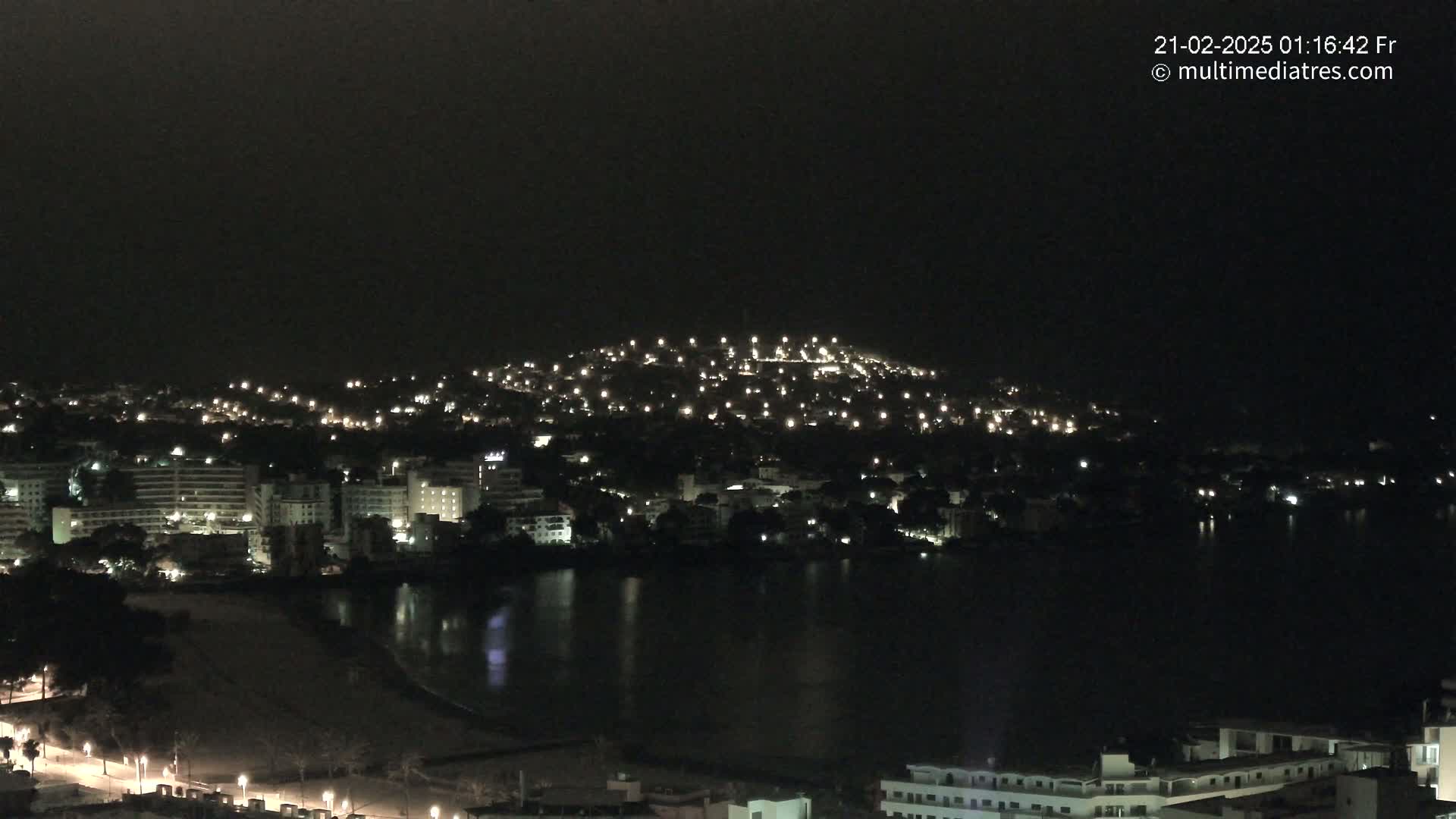 A nighttime view of a city on a hill next to a dark body of water, illuminated by numerous lights.